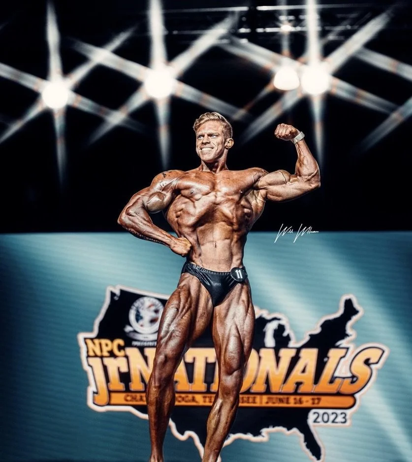 Bodybuilder posing on stage at NPC Junior Nationals with event banner in background, bright stage lights above.
