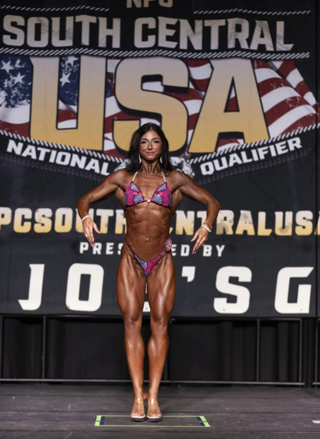 A female bodybuilder posing on stage during a USA bodybuilding competition, wearing a pink and purple bikini and clear high heels.