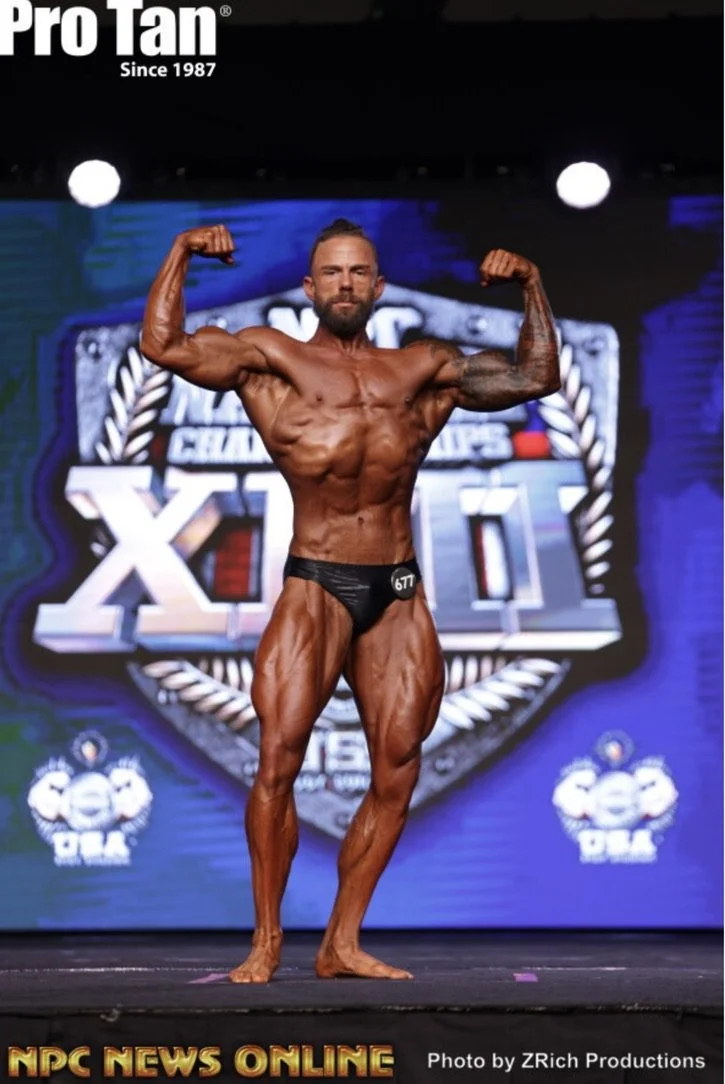 Bodybuilder flexing muscles on stage with Pro Tan and NPC News Online logos.