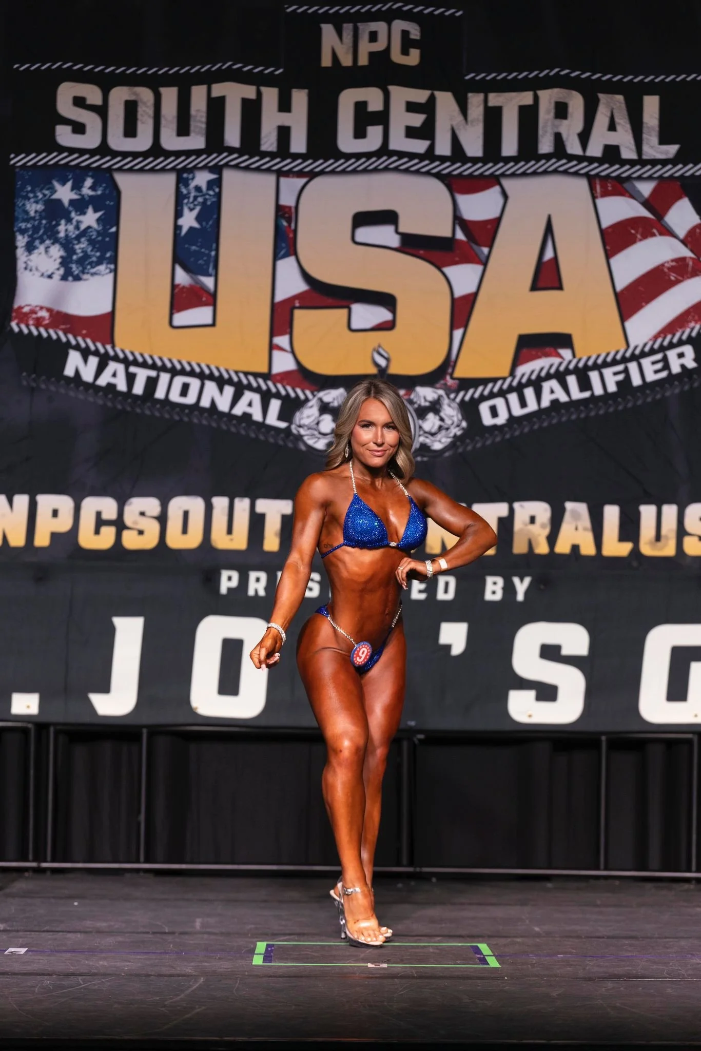 A woman in a blue bikini standing on a stage with a large backdrop that says 'NPC South Central USA National Qualifier' and 'Presented by J.O.S.O.' in the background.
