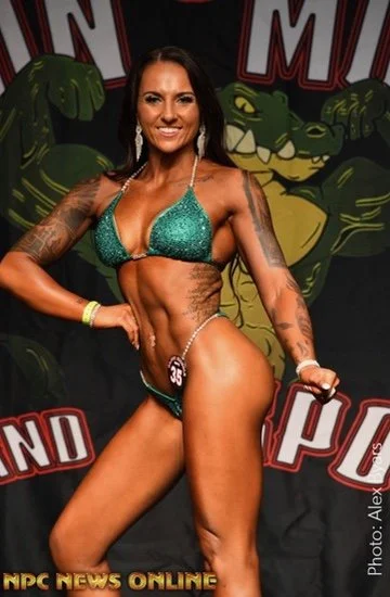 Bodybuilder posing in a green bikini on stage with a reptile-themed backdrop at a bodybuilding competition.