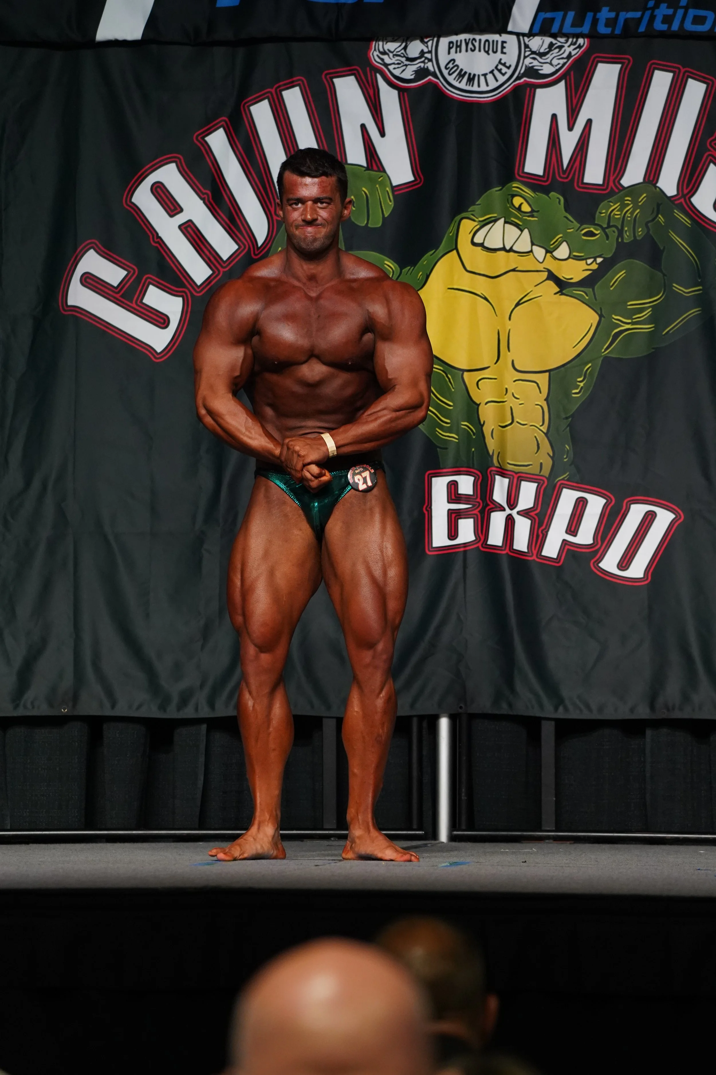 A male bodybuilder on stage in a bodybuilding competition, posing with flexed muscles, wearing a dark green speedo, in front of a black curtain with a logo featuring a muscular crocodile and the text "Chicken Mud Expo."