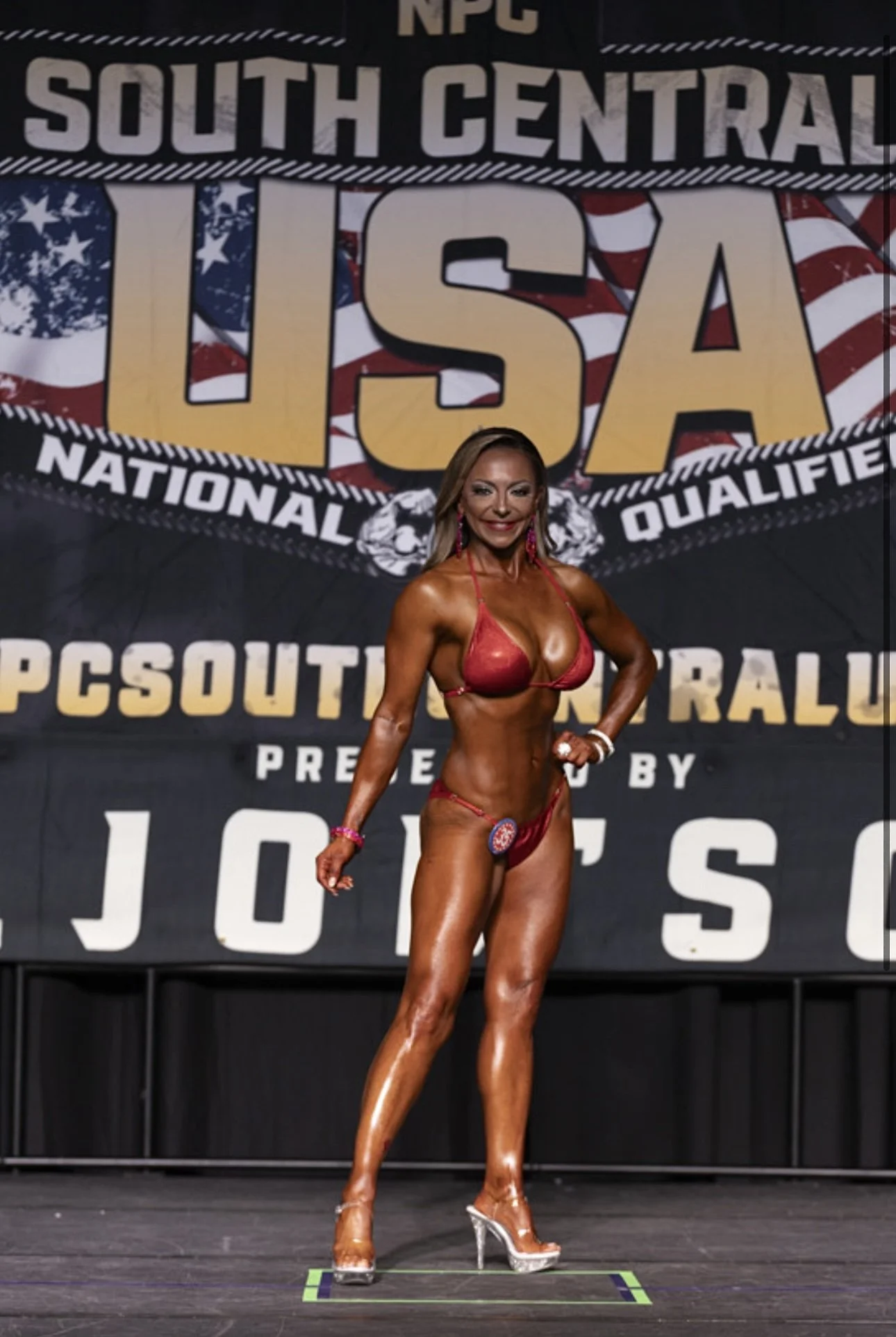 A female bodybuilder in a red bikini and clear high heels standing on stage at a fitness competition, with a large USA-themed banner in the background.
