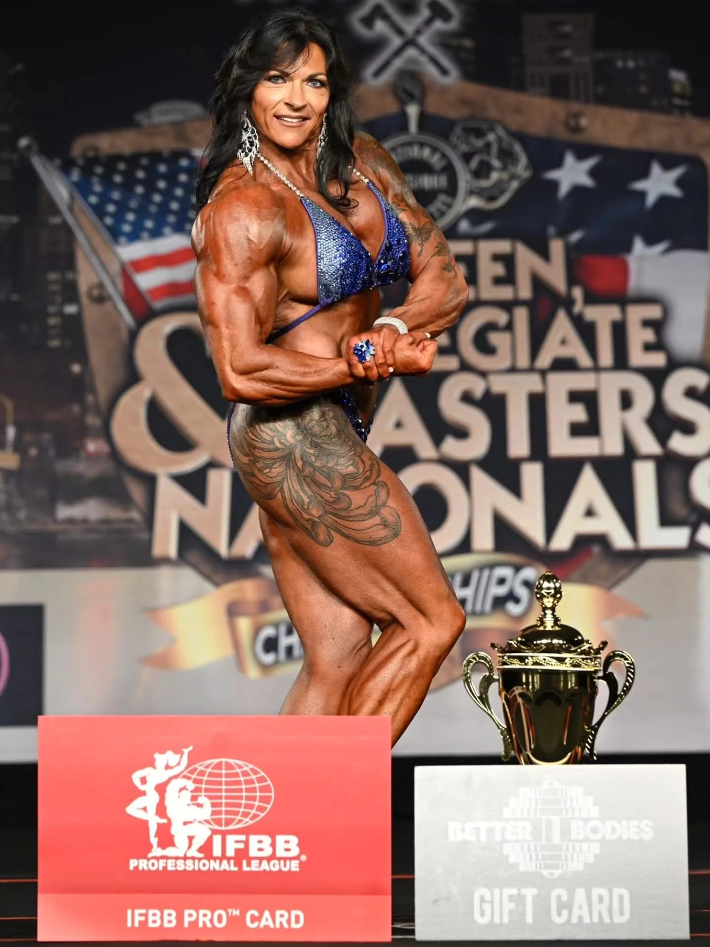 Female bodybuilder on stage at a competition, flexing her arm. She is wearing a blue bikini and jewelry, with tattoos on her leg and arm. Behind her is a large backdrop with the words "Golden Gladiator Masters Nationals." A gold trophy is on a podium