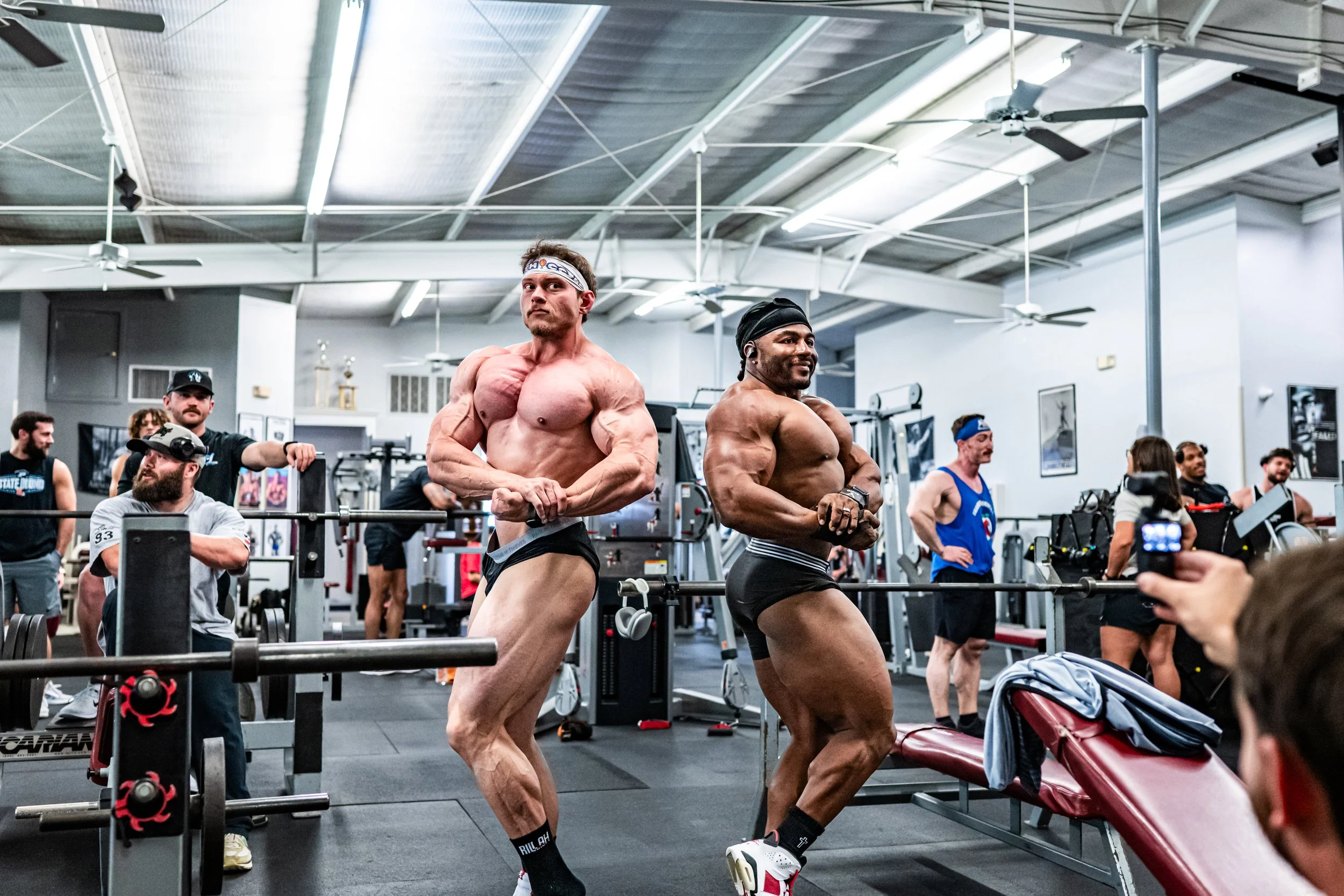 Two muscular men with minimal clothing, wearing headbands and posing at a gym during a bodybuilding competition or photoshoot, surrounded by gym equipment and spectators.