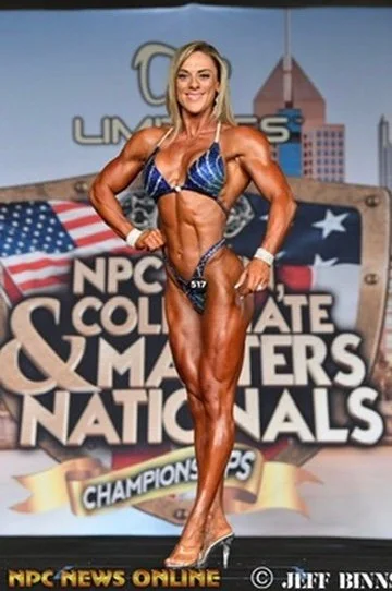 A female bodybuilder poses on stage at the NPC Collegiate & Masters Nationals Championships. She's wearing a blue bikini and high heels, displaying a muscular physique. The background shows event branding and an American flag.