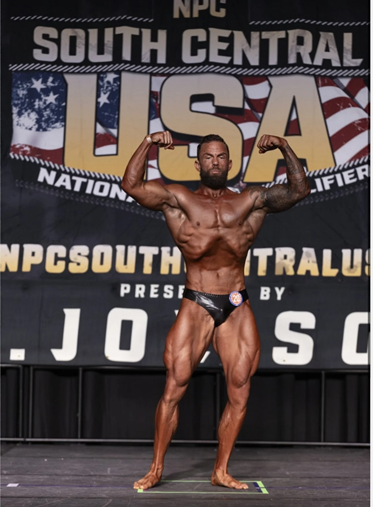 A male bodybuilder on stage in a bodybuilding competition, flexing his muscles, wearing black posing trunks, with a backdrop reading 'South Central USA' and 'NPC South.'