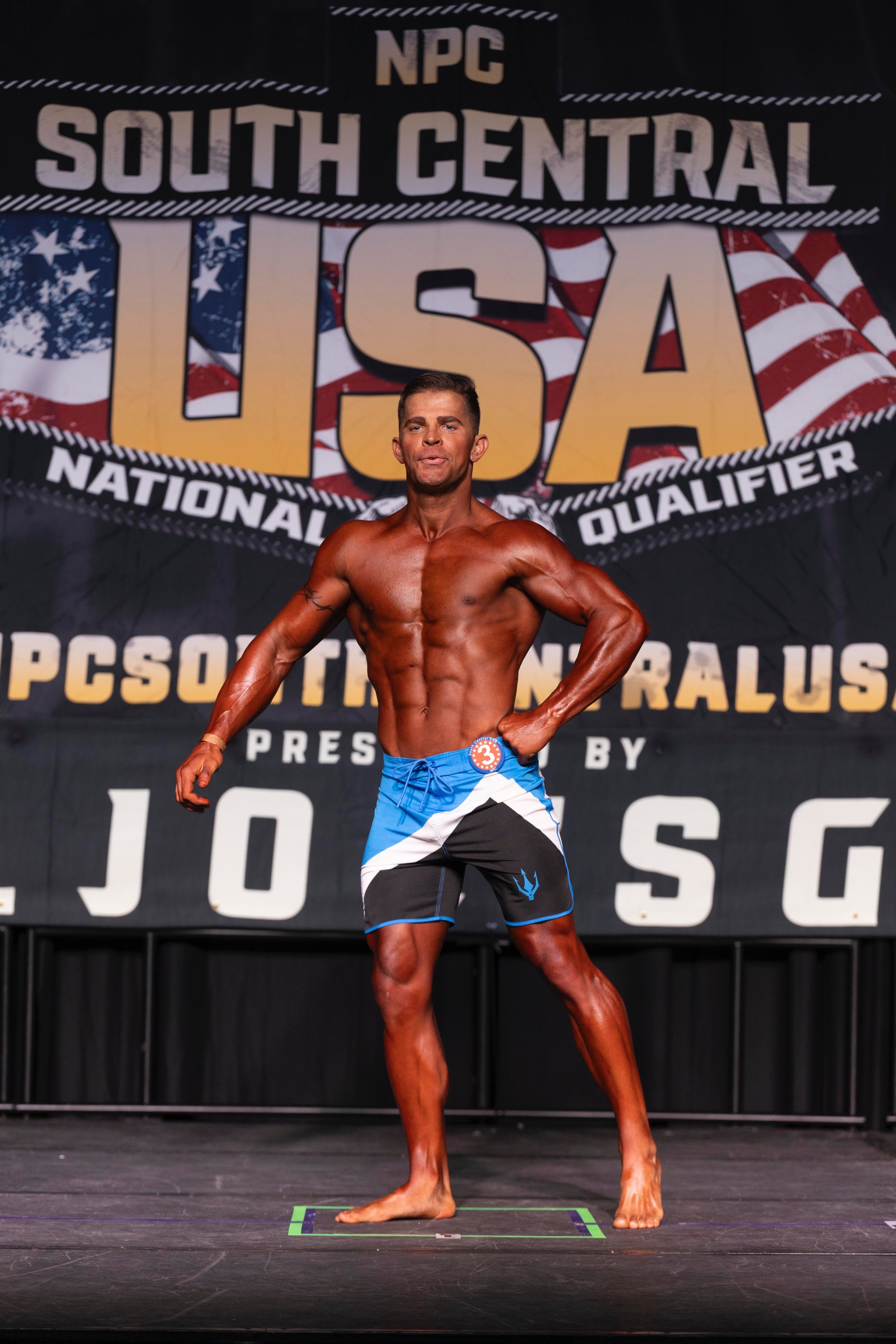 A muscular male bodybuilder on stage at the NPC South Central USA bodybuilding competition, posing in front of a banner.