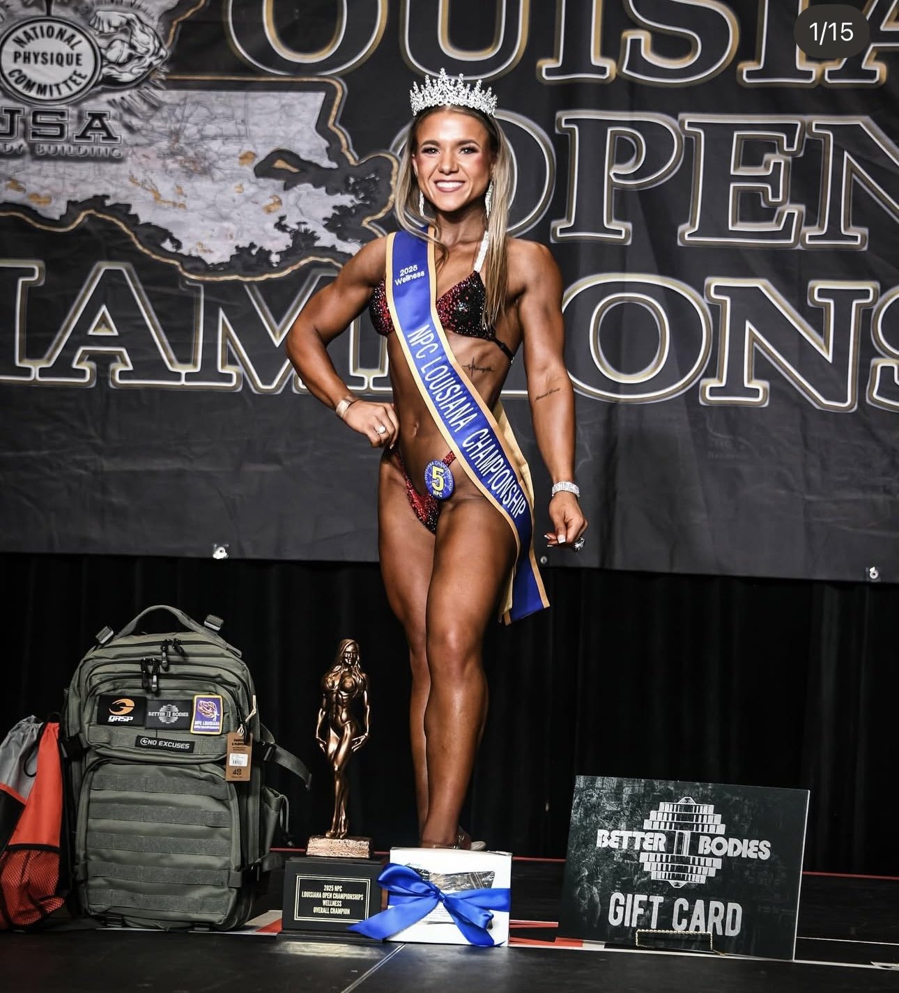 A female bodybuilder on stage wearing a crown and a sash that reads 'NPC Louisiana Championship 2023.' She is smiling and posing with her right hand on her hip. On the stage, there is a trophy, a gift card, and some gear.