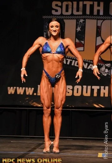 Female bodybuilder posing on stage in a blue bikini with number 27, during a competition, showcasing muscle definition and symmetry. Event banner visible in the background.