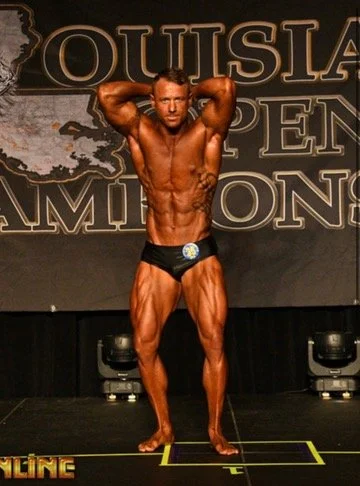 Bodybuilder posing on stage at the Louisiana Open Championships with a muscular physique, wearing black trunks.