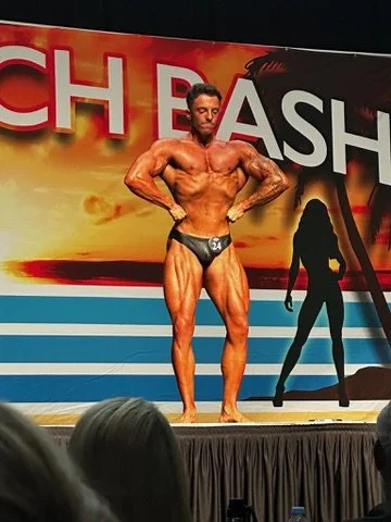 Bodybuilder posing on stage at a competition with a beach-themed background.