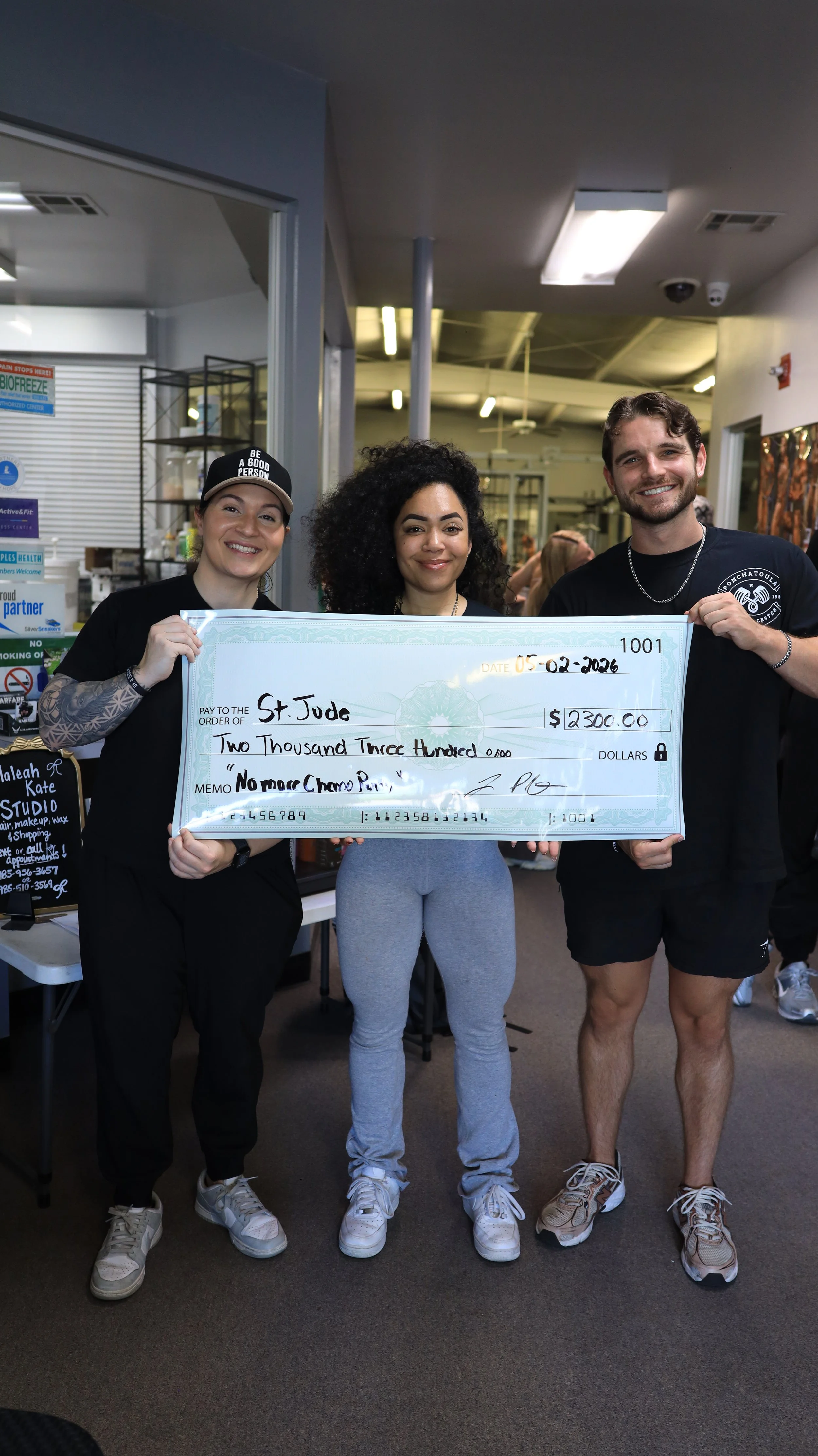 Three people holding a large check, smiling, inside a store or indoor setting. The check is made out to St. Jude for $2,300, and the date is February 5, 2026.