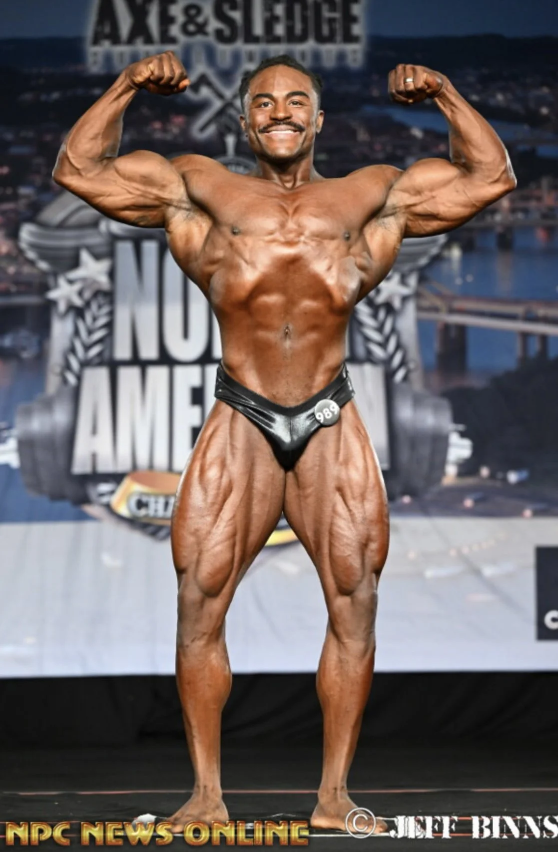 A male bodybuilder with dark hair and a mustache poses with flexed arms on stage at a bodybuilding competition, wearing a black posing suit with the number 989, in front of a backdrop with the text 'Axe & Sledge' and 'NPC News Online' at the bottom.