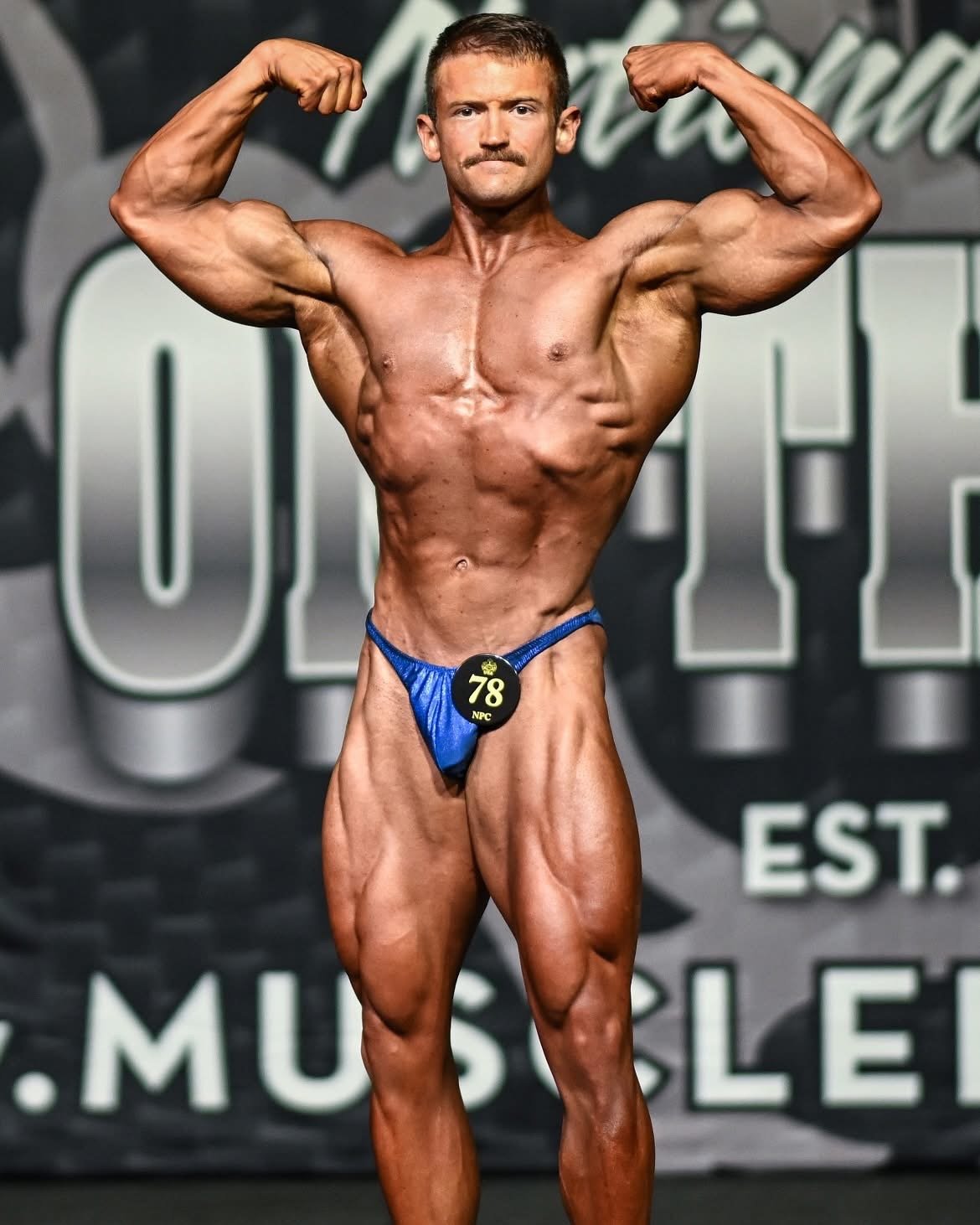 A male bodybuilder in a blue posing suit flexing his muscles on stage during a competition.