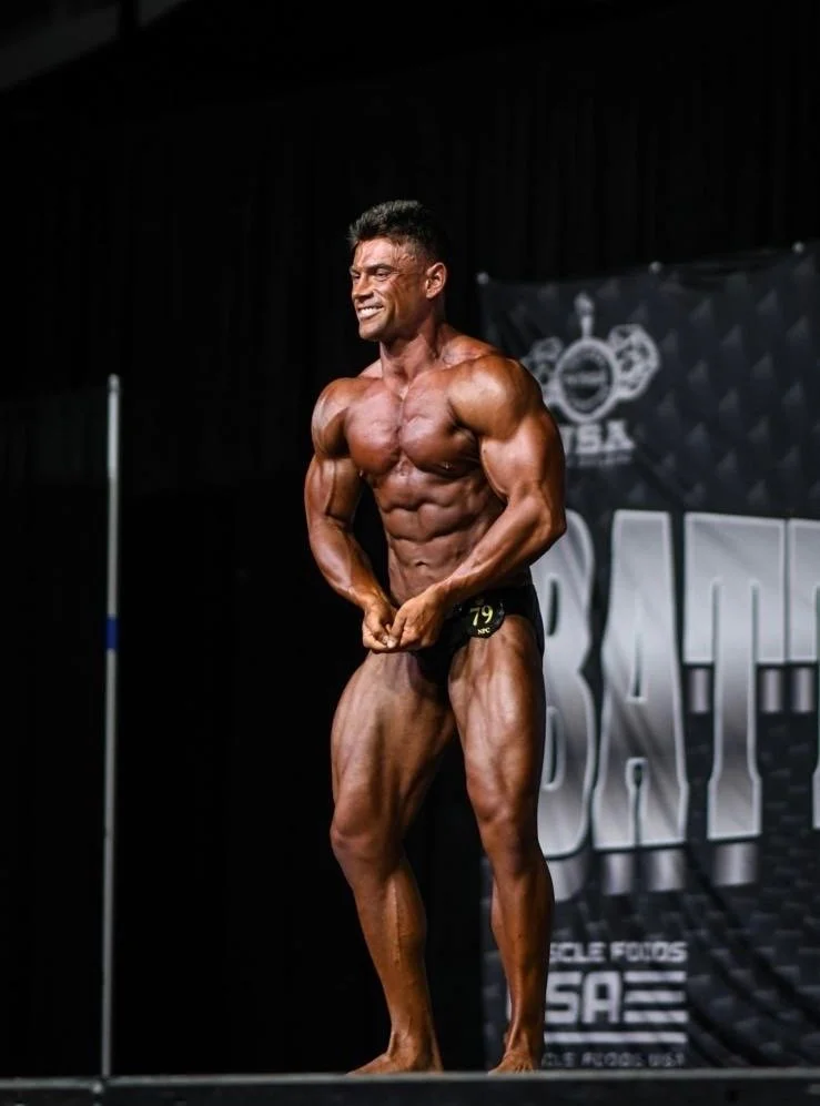 Male bodybuilder on stage during competition, smiling, flexing muscles, wearing black posing trunks, black backdrop, fitness branding visible.