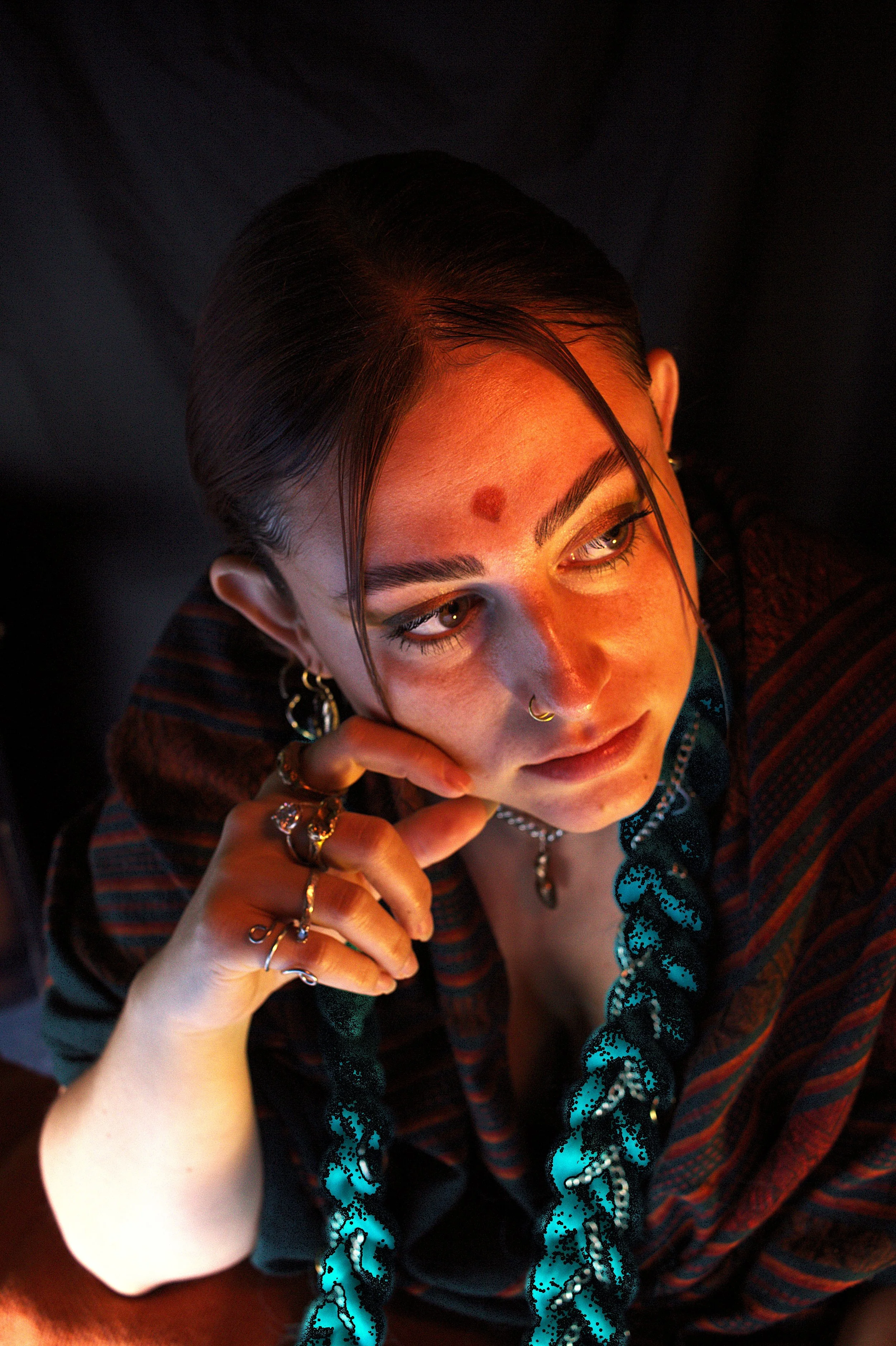 A young woman with dark hair styled in braids, wearing multiple rings, earrings, a nose ring, and a necklace, resting her chin on her hand and looking to the side against a dark background.