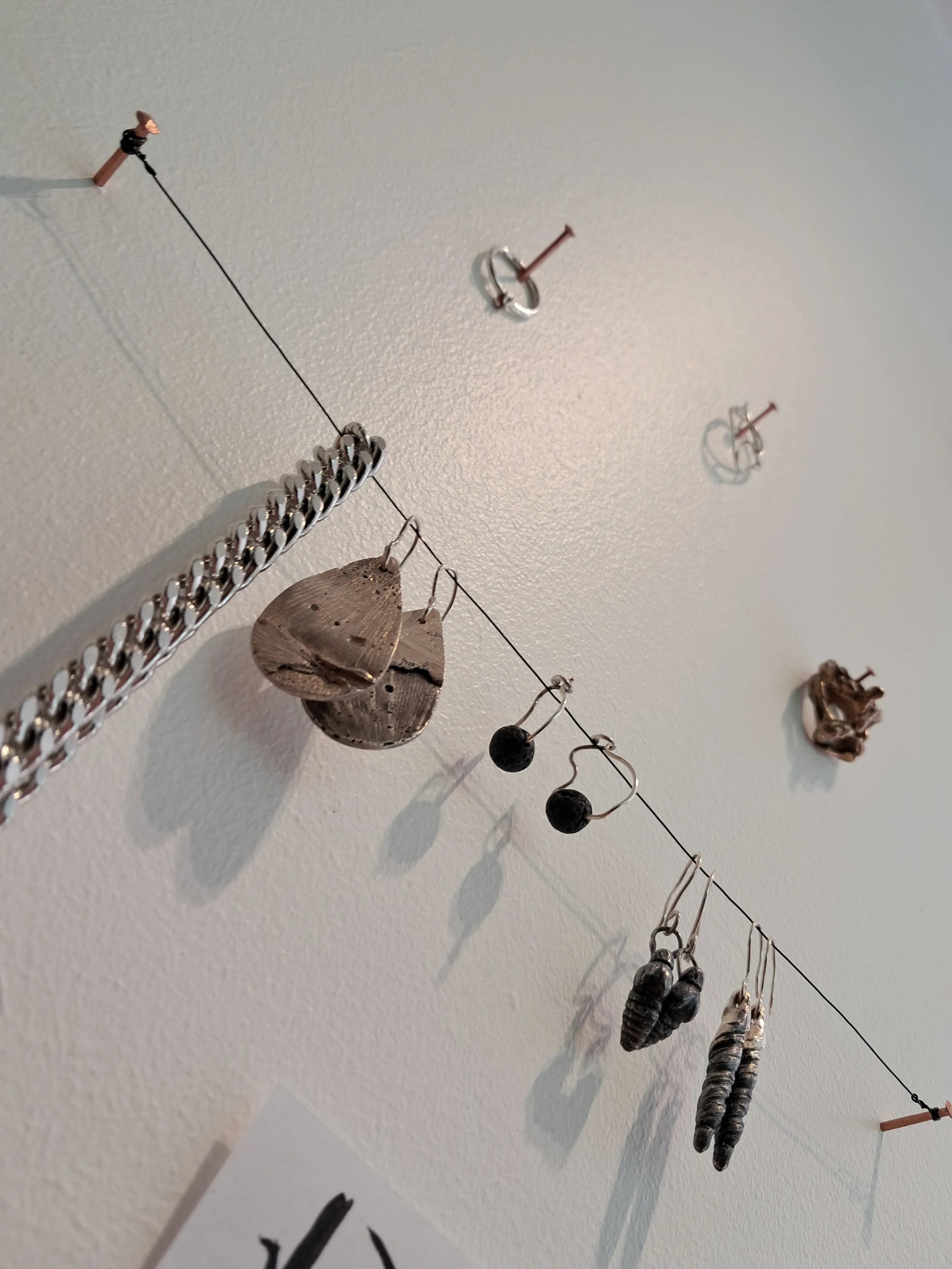 Various jewelry pieces hanging from a thin wire on a white wall, including earrings, necklaces, and decorative items with shadows cast on the wall.