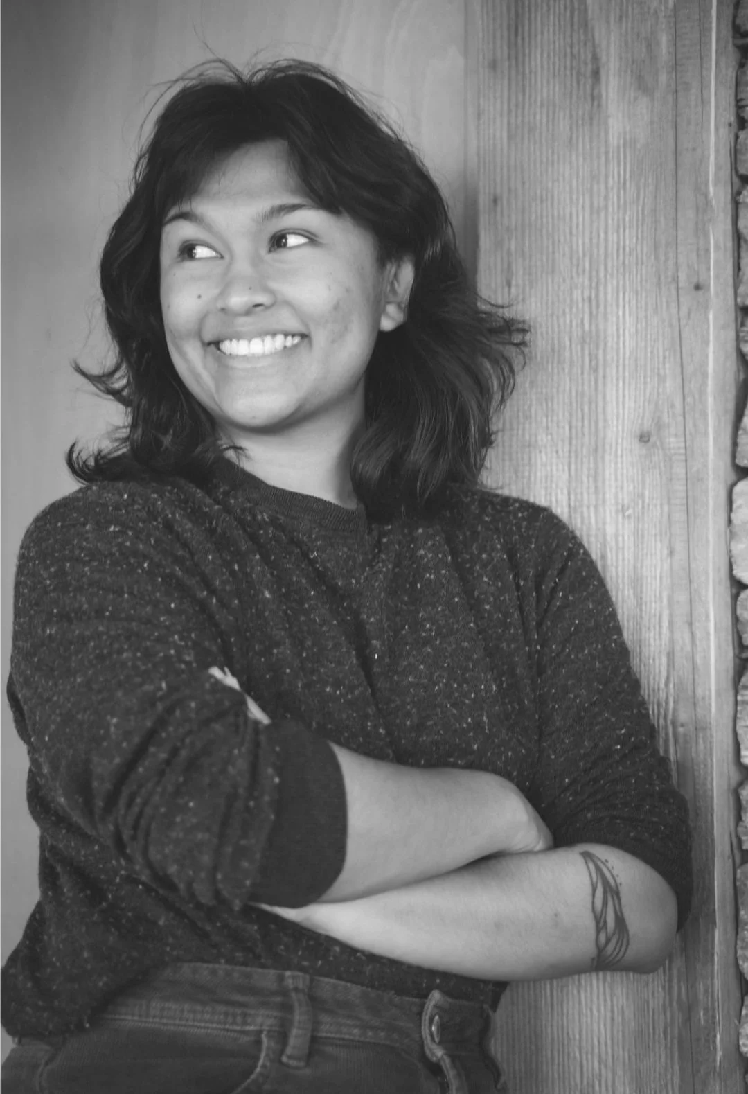 Black and white photograph of a young woman with shoulder-length hair smiling and looking to her left, standing with arms crossed against a wooden wall.