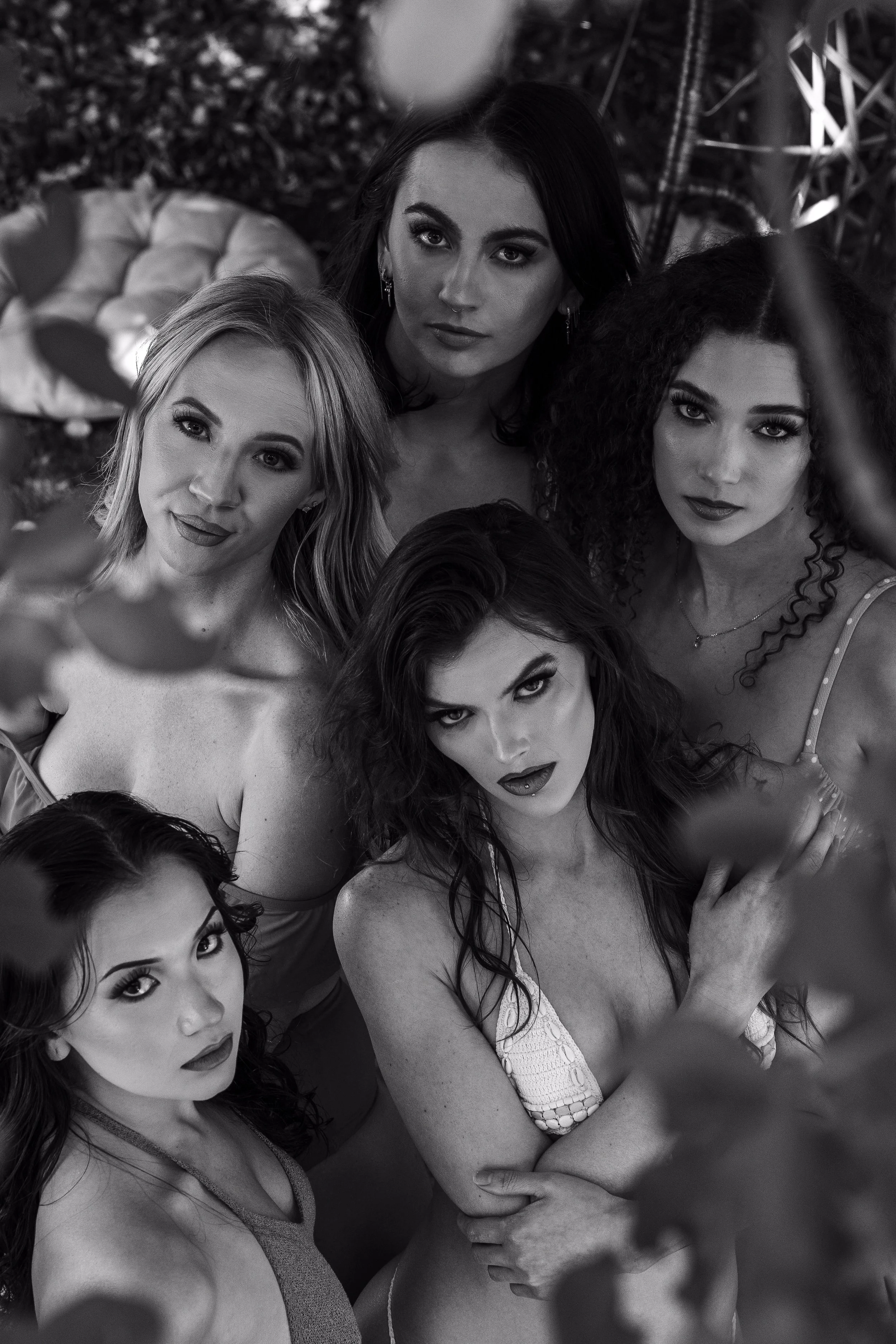 Group of five women taking a selfie outdoors, looking up at the camera, with leaves and a cushioned seat in the background, in black and white.