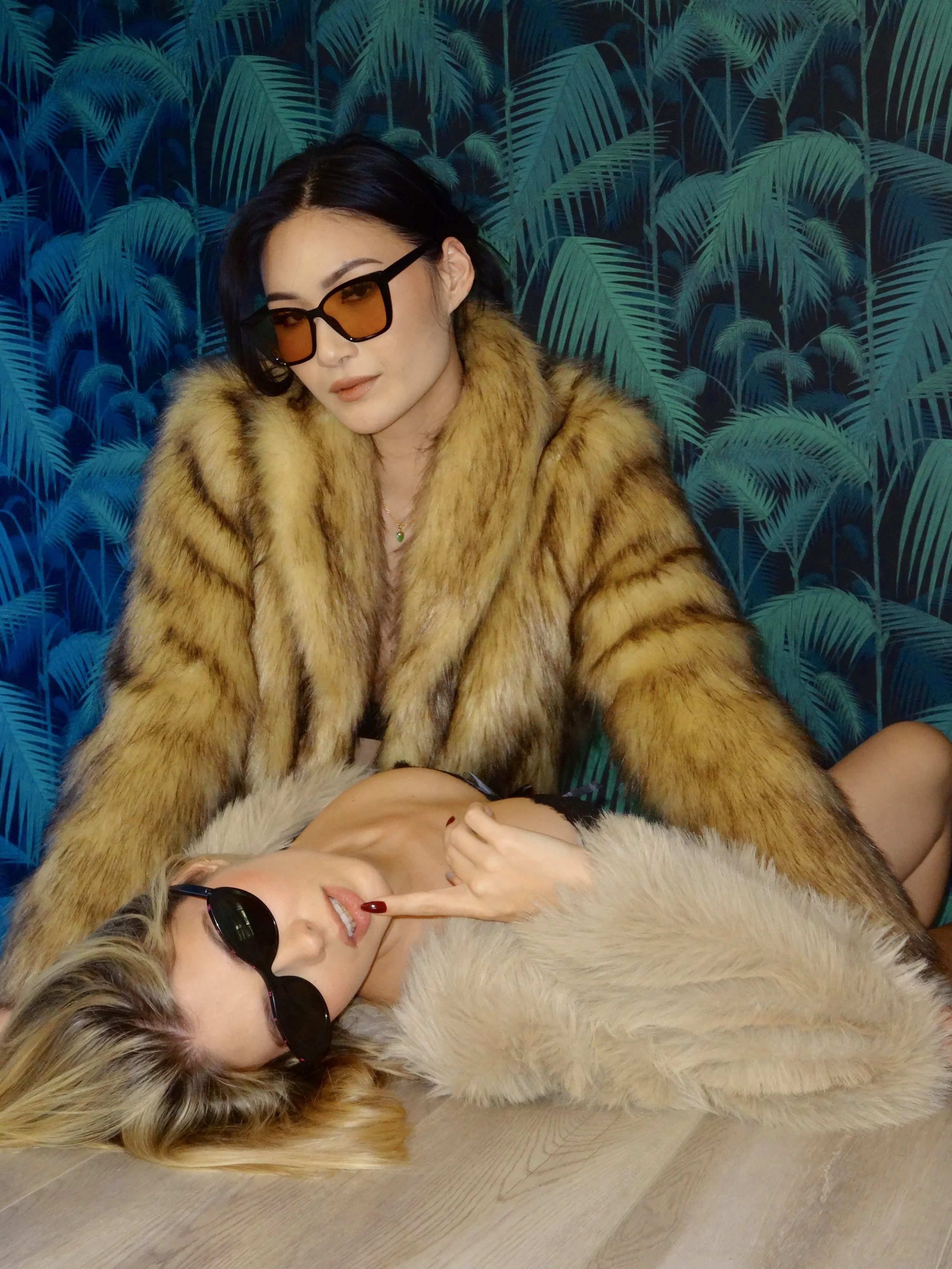 Two women wearing fur coats and sunglasses pose against a tropical palm wallpaper background. One is lying on the floor with black sunglasses, the other is sitting behind her with orange-tinted glasses.