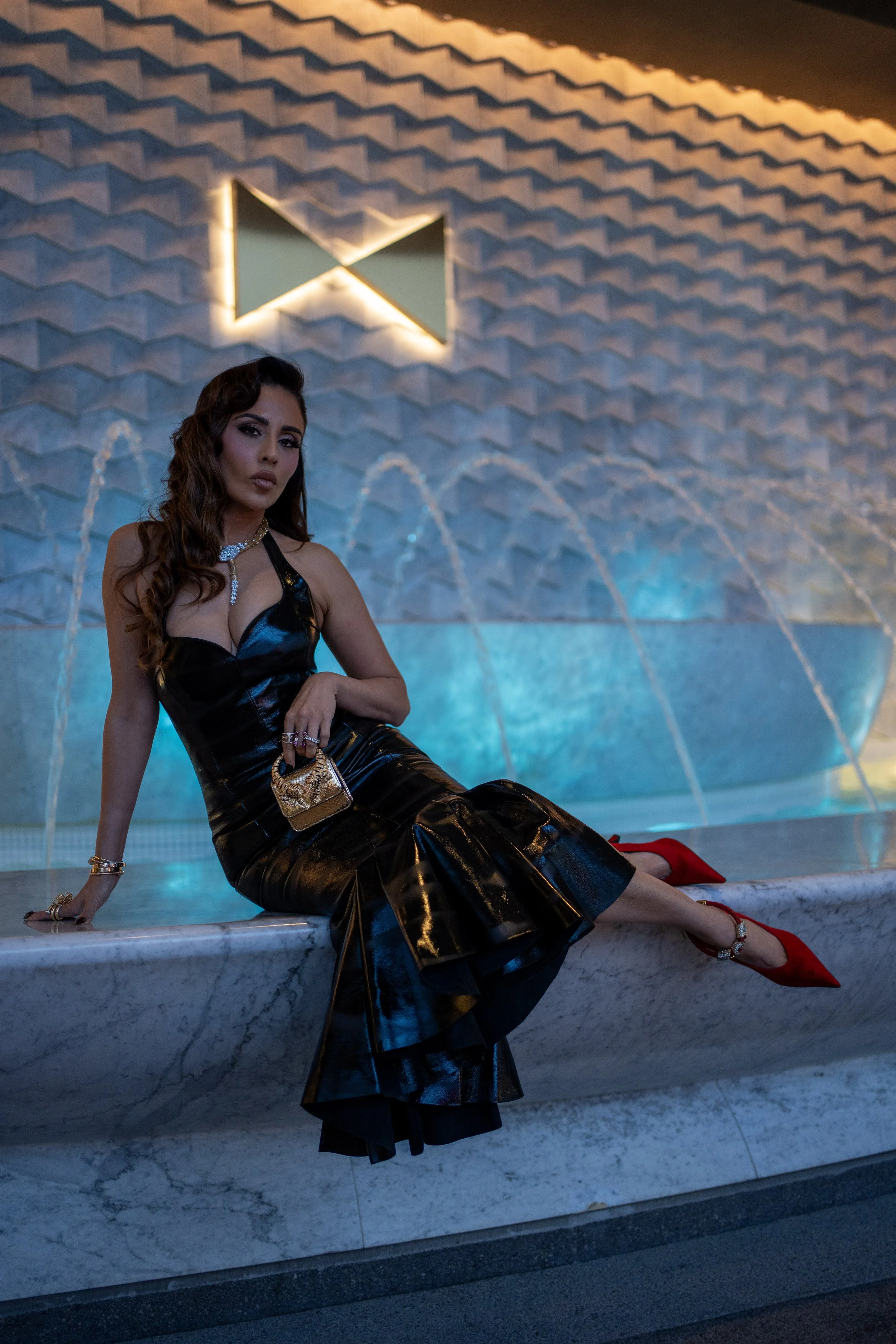 A woman in a black shiny dress sitting on a marble ledge beside a fountain with arched streams of water, in front of a textured wall with backlit geometric shapes.