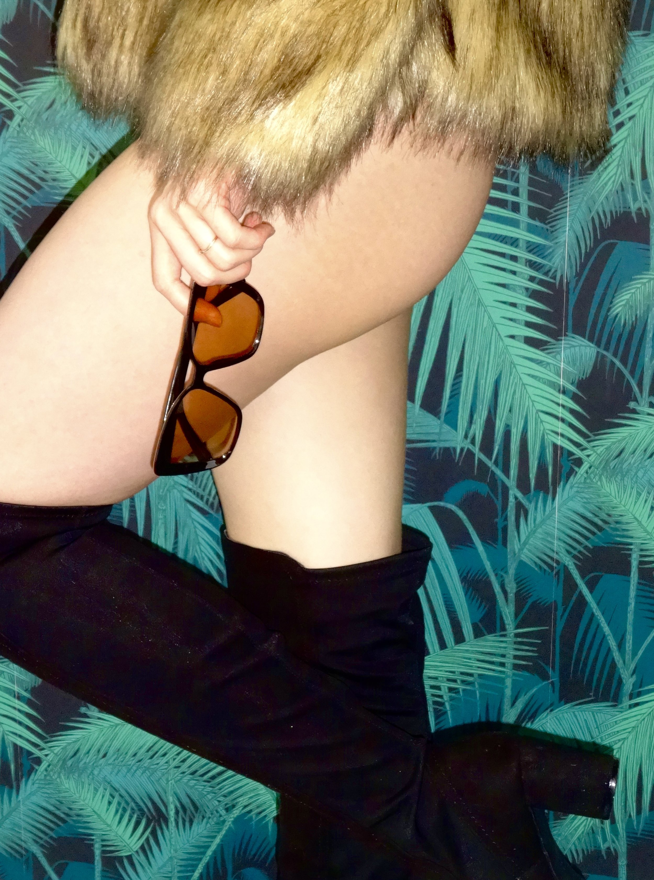 A woman in thigh-high black boots, holding orange-tinted sunglasses, with a leopard-print coat, against a background of tropical leaves.