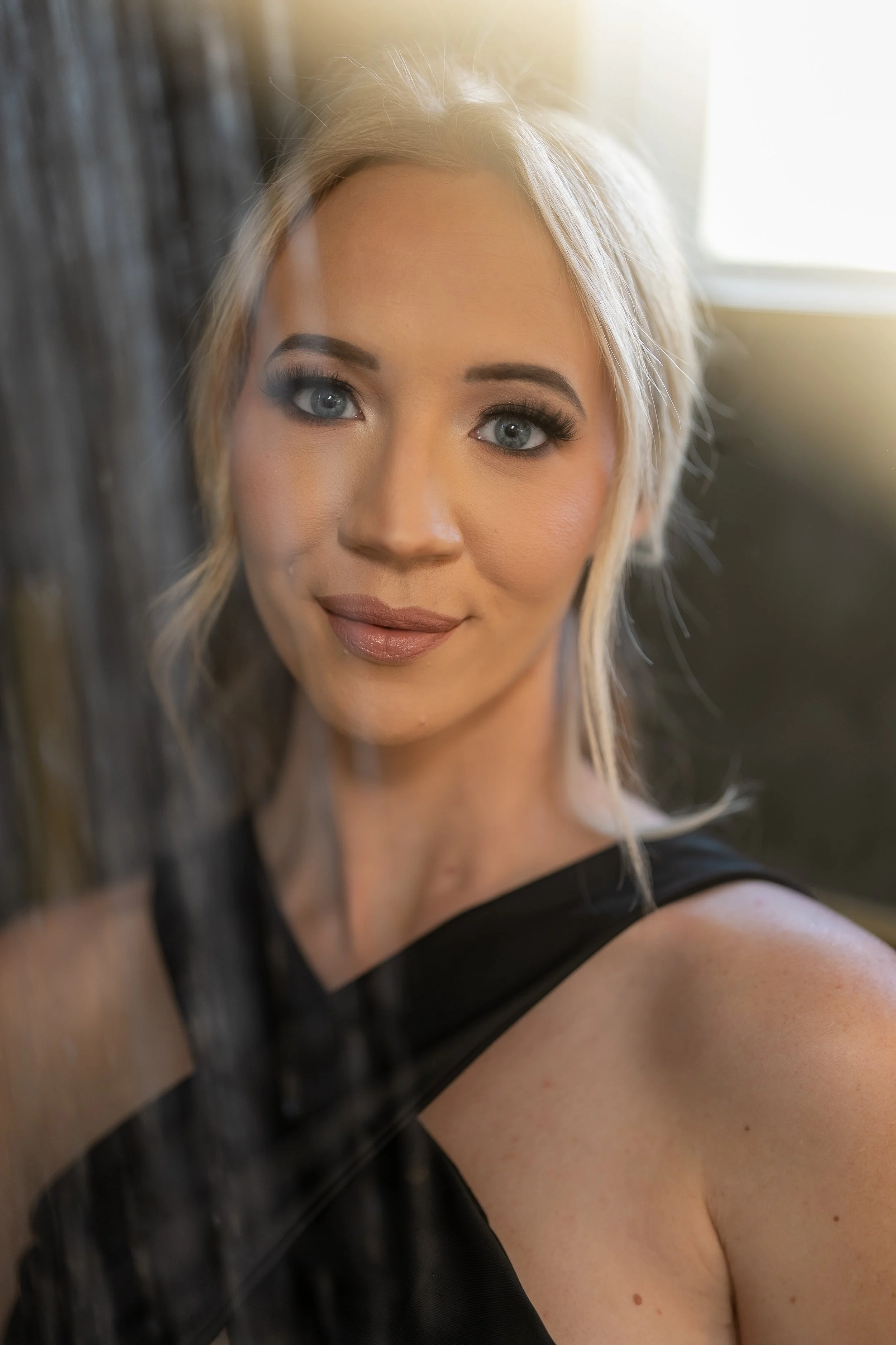 Close-up portrait of a blonde woman with blue eyes, wearing black clothing, standing near a wooden surface with sunlight behind her.