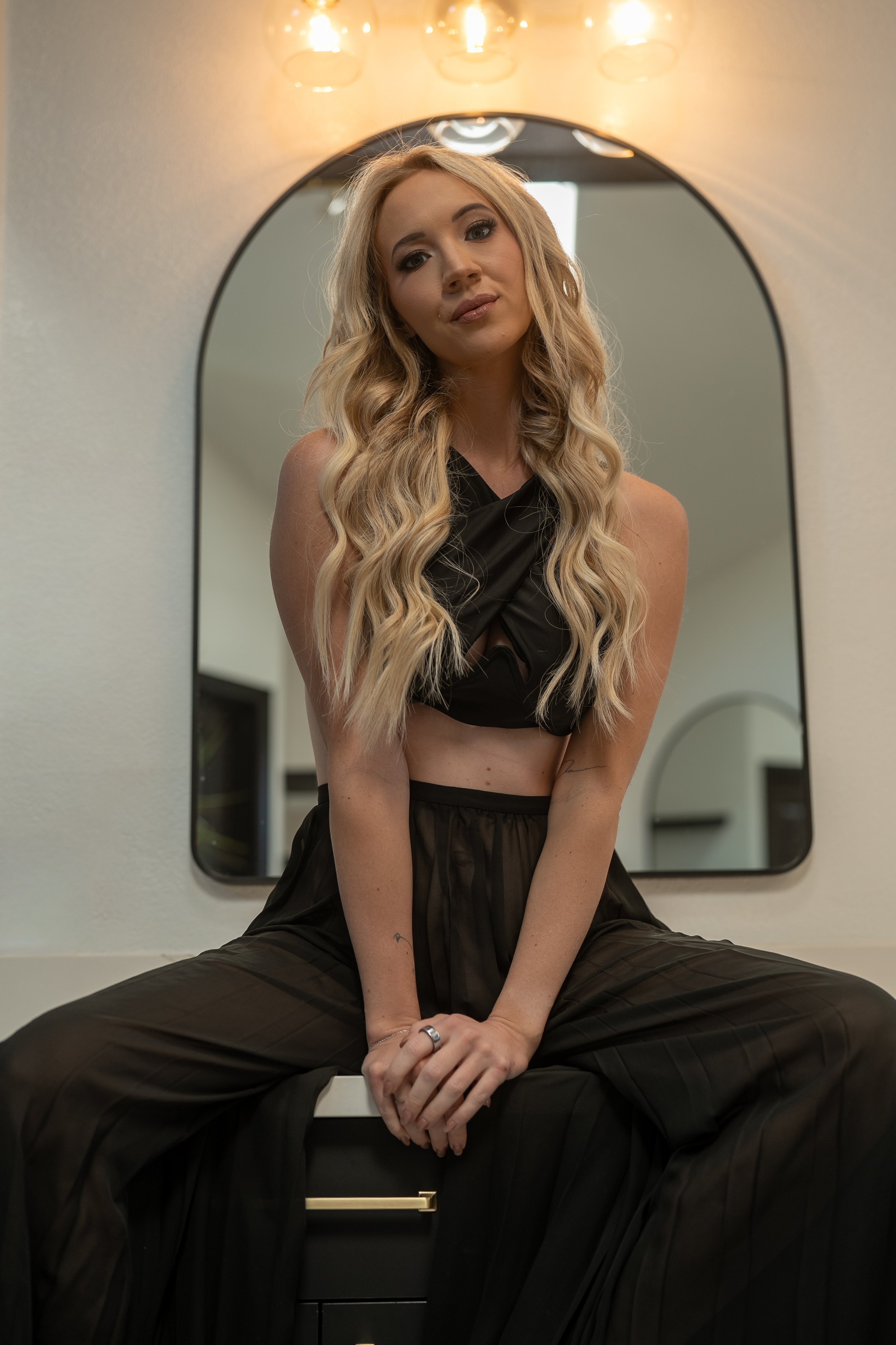 A young woman with long, wavy blonde hair sits on a vanity table with her legs spread apart, wearing a black sleeveless top and matching wide-legged pants, in front of a mirror with warm lighting.