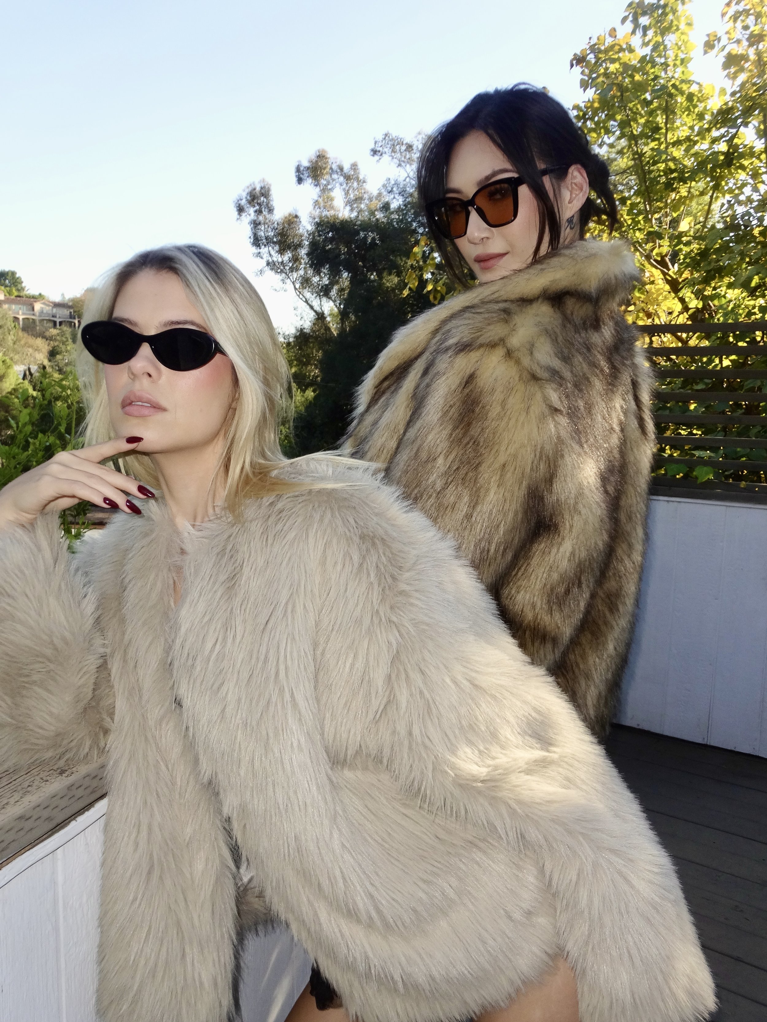 Two women wearing fur coats and sunglasses posing outdoors with trees and a fence in the background.
