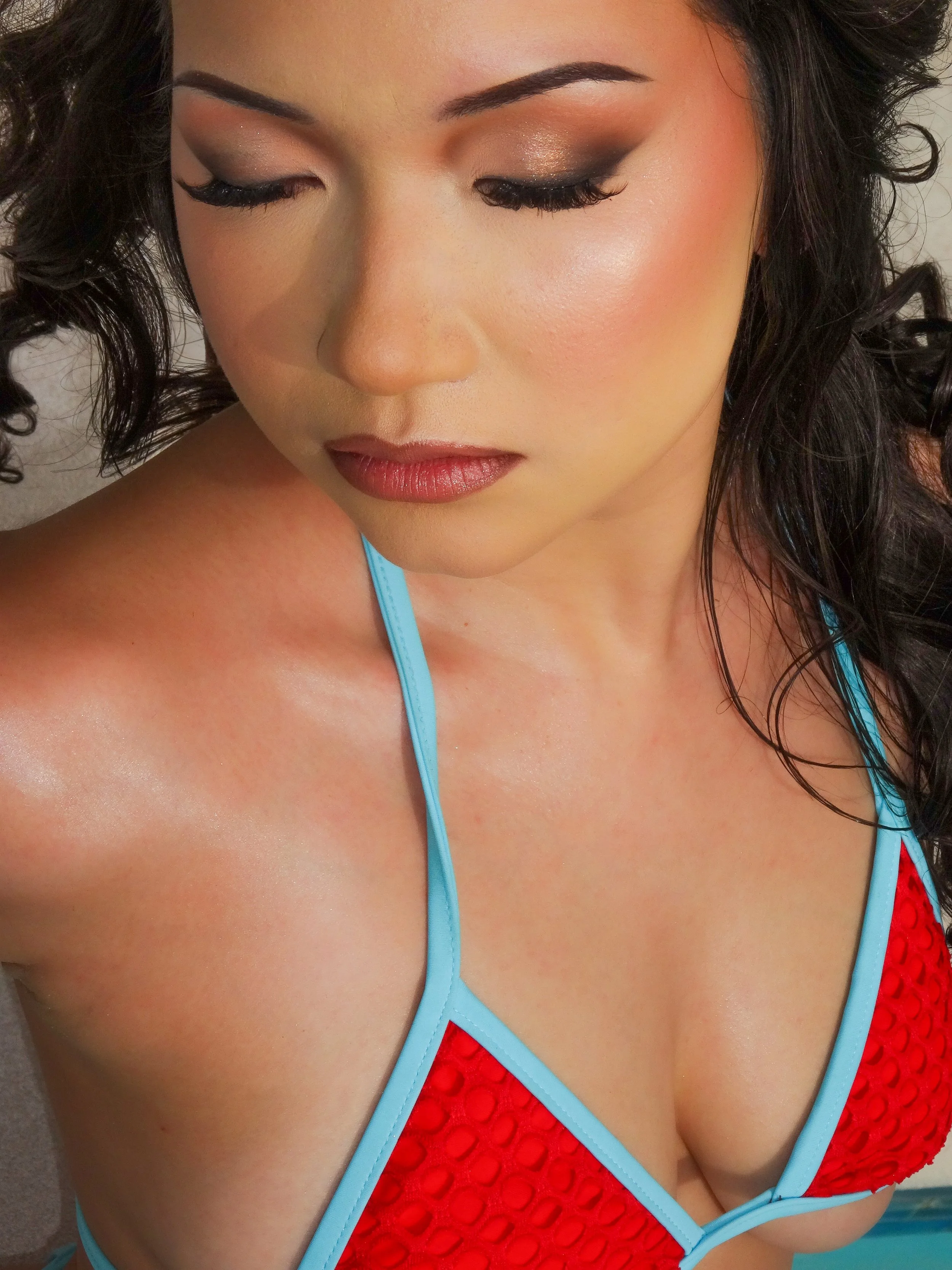A woman with dark hair, closed eyes with makeup, and wearing a red bikini top with blue trim, looking downward.