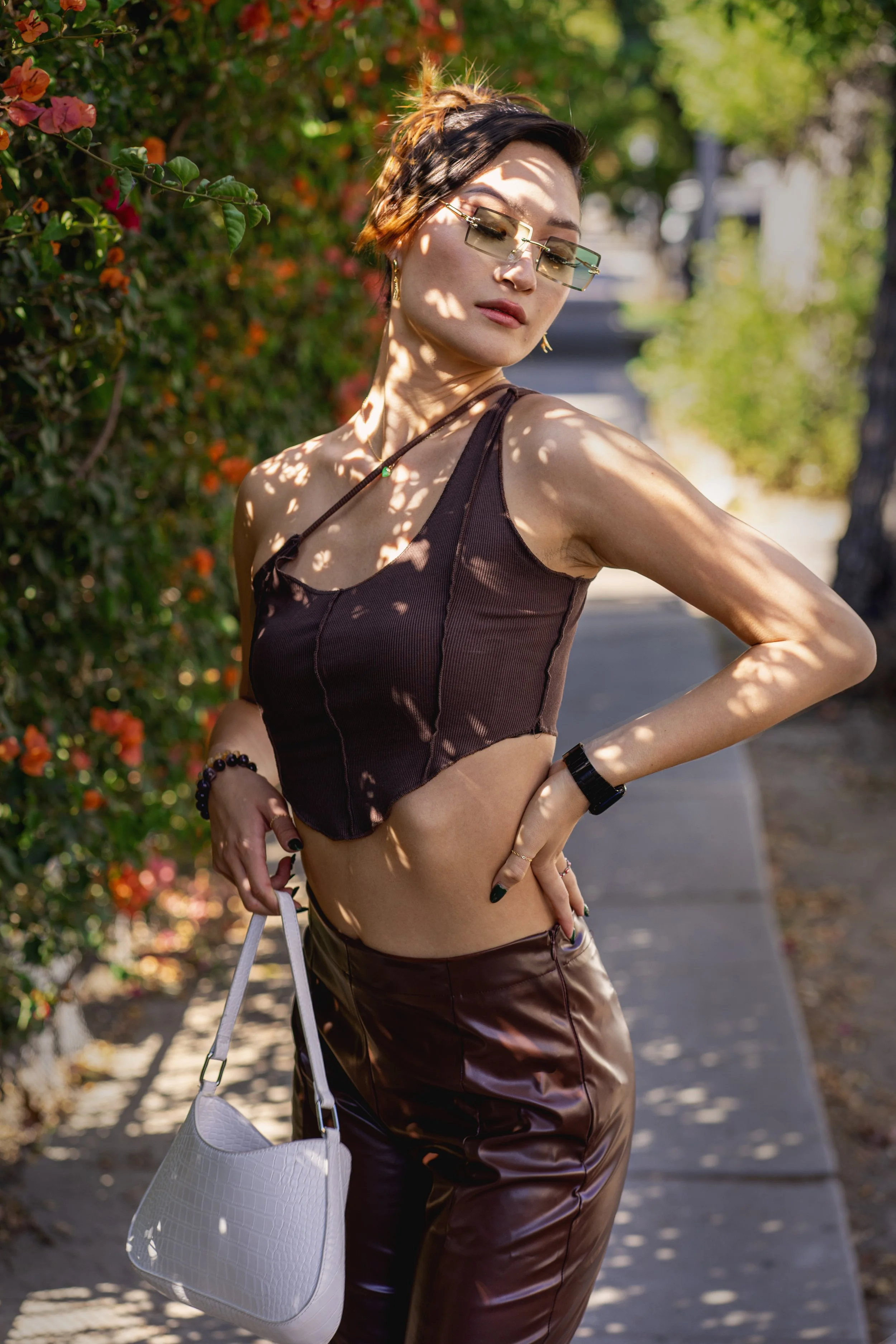 A woman standing outdoors on a sidewalk with her hand on her hip, wearing sunglasses, a dark sleeveless top, and shiny dark brown pants, carrying a white purse, with sunlight filtering through trees casting shadows on her.