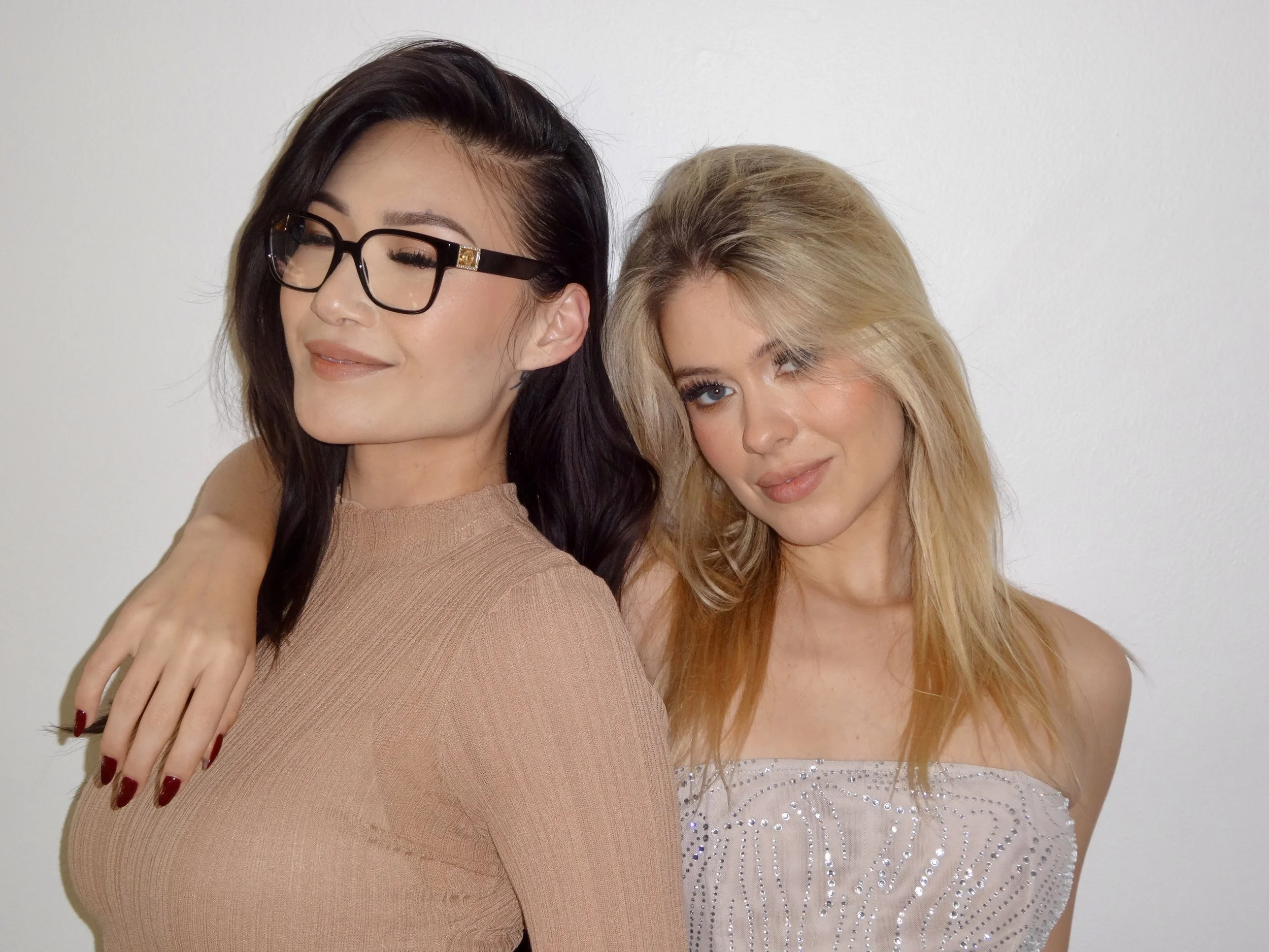 Two women standing close together with one woman having her arm around the other, both smiling and posing for a photo against a plain white background.