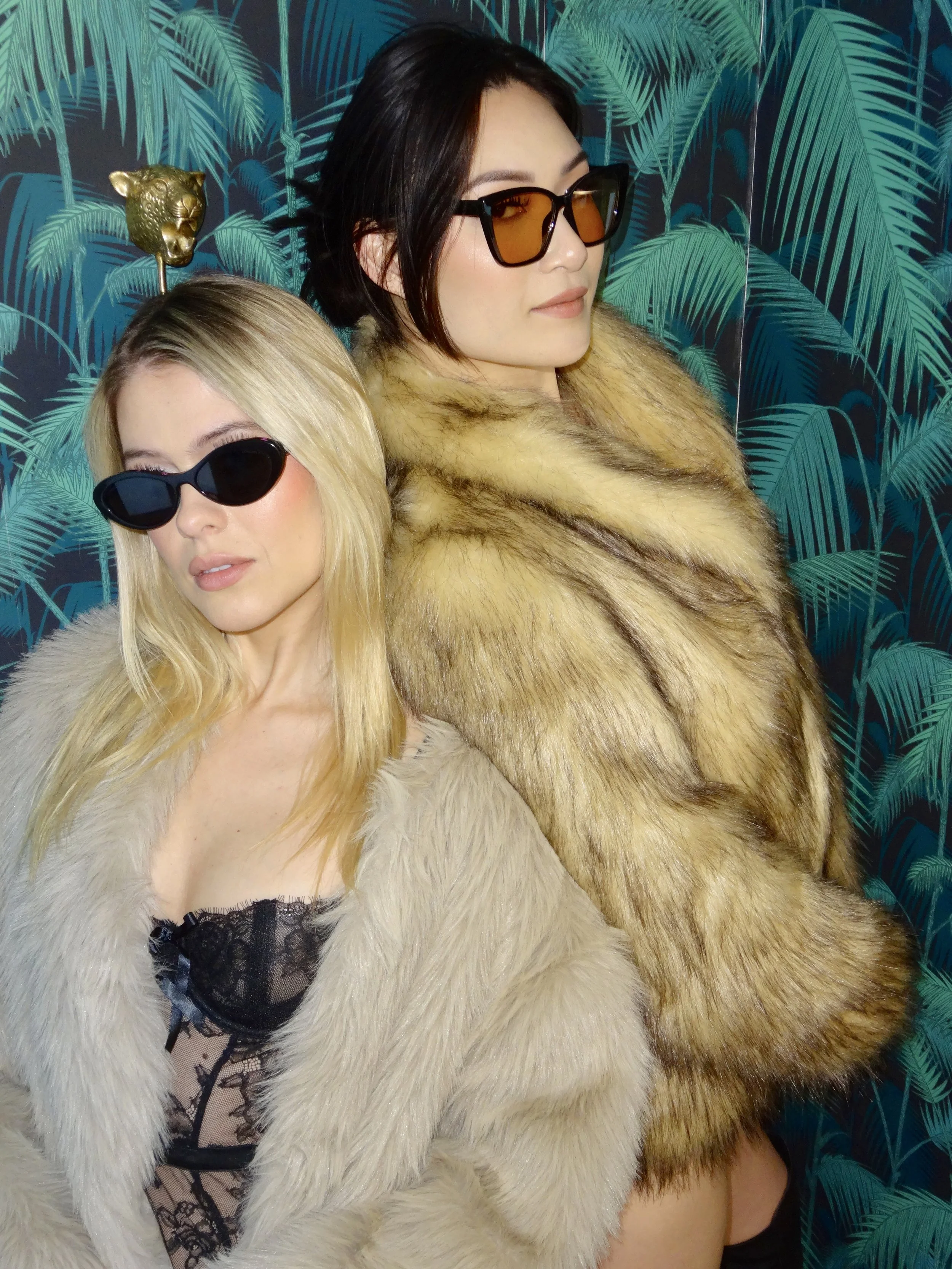 Two women wearing fur coats and sunglasses posing against a leafy, tropical-patterned background.