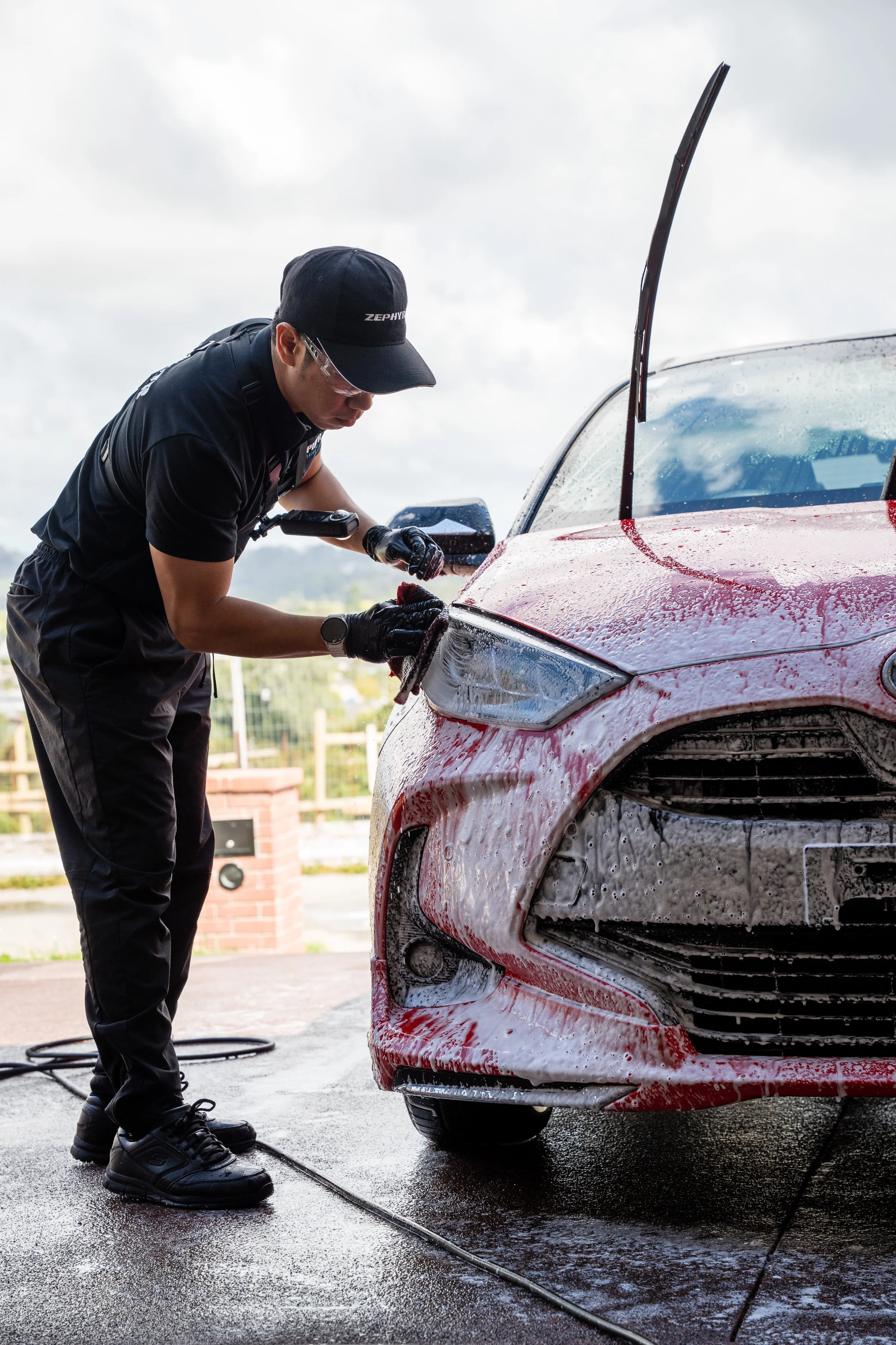 Person washing the front of a red car with soap suds during daytime.