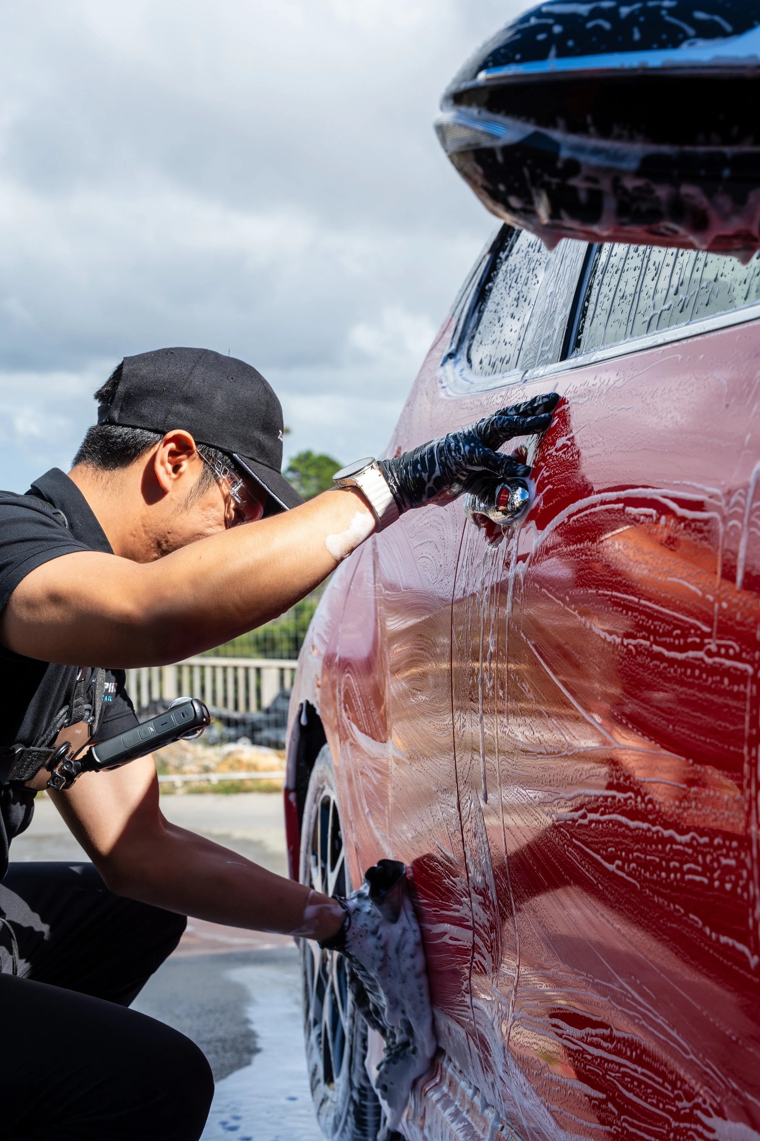 A person wearing gloves and a cap is washing a red car with soap and a sponge outdoors on a cloudy day.