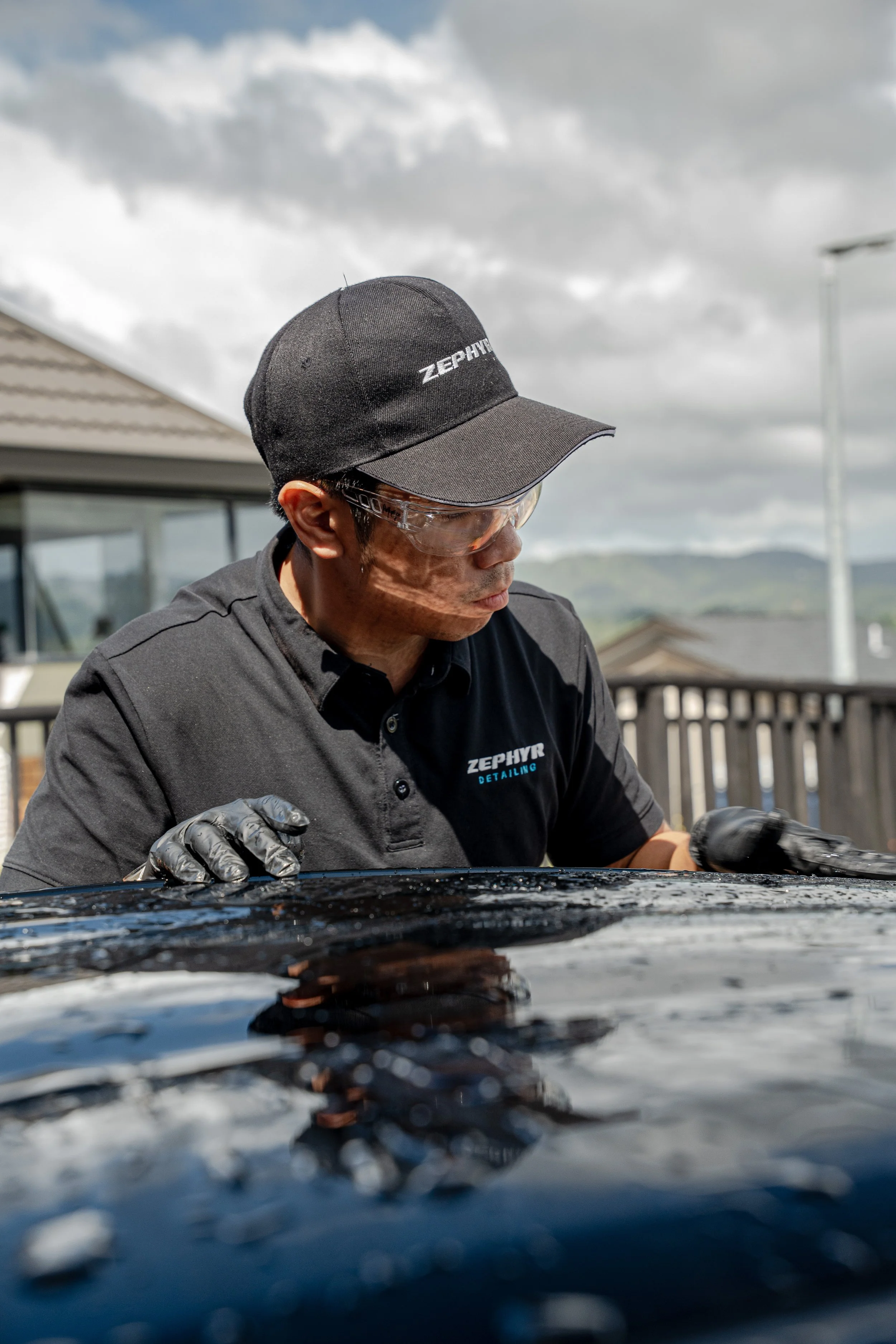 A man wearing safety glasses, a black cap, black gloves, and a black polo shirt with the words 'ZephyR Detailing' on it is polishing a car outdoors under a cloudy sky.