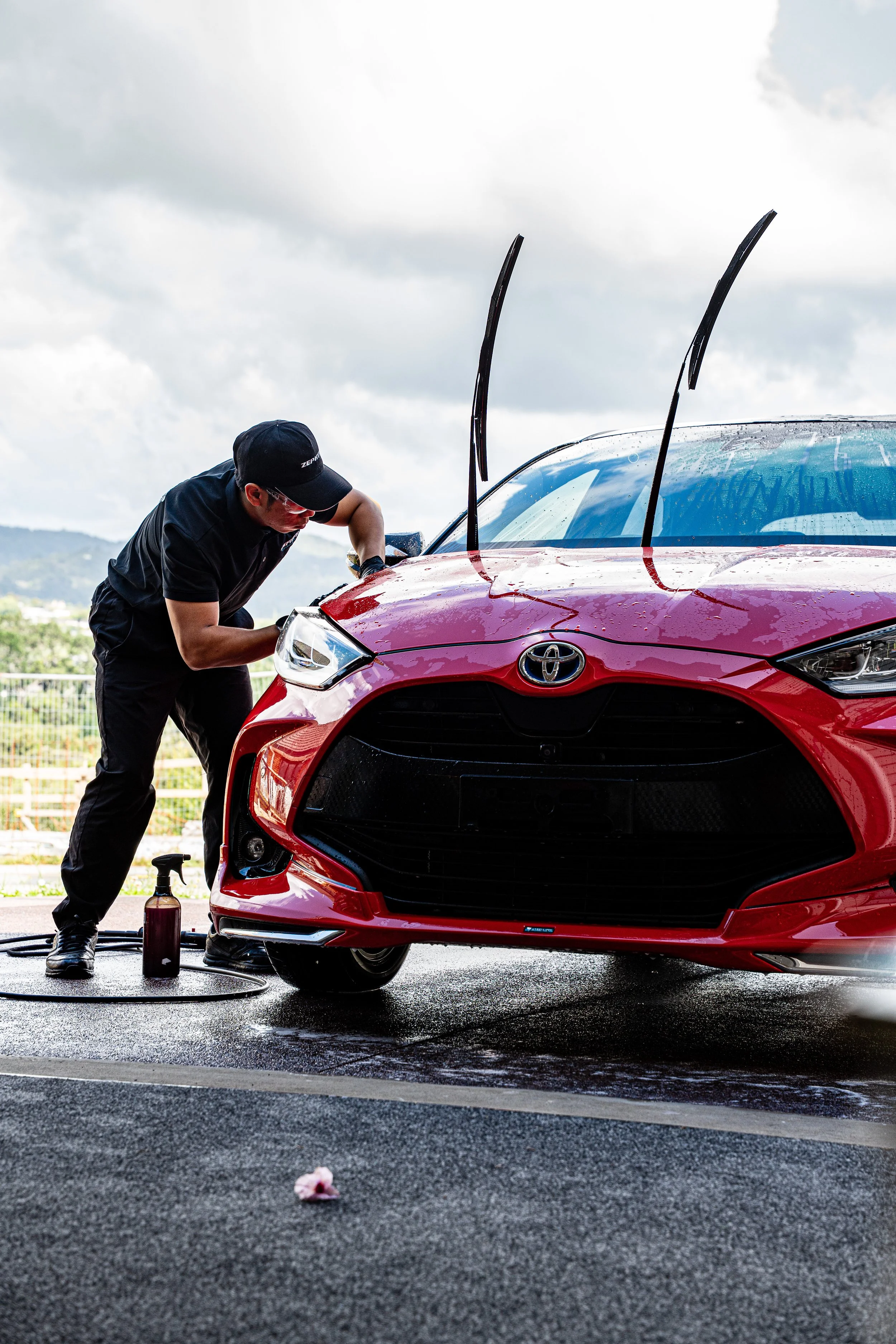 A man cleaning a red Toyota race car with water and a spray bottle at a track, with hills and cloudy sky in the background.