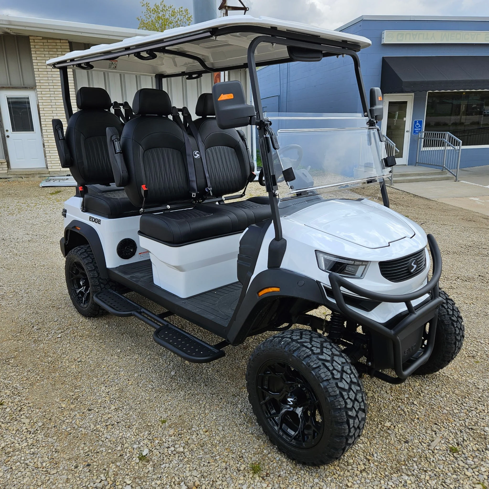 A white golf cart with black seats and a clear windshield parked outside a building with blue and beige walls.