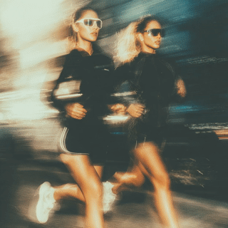 Two women running side by side at night, wearing athletic clothing and sunglasses, with a motion blur effect.