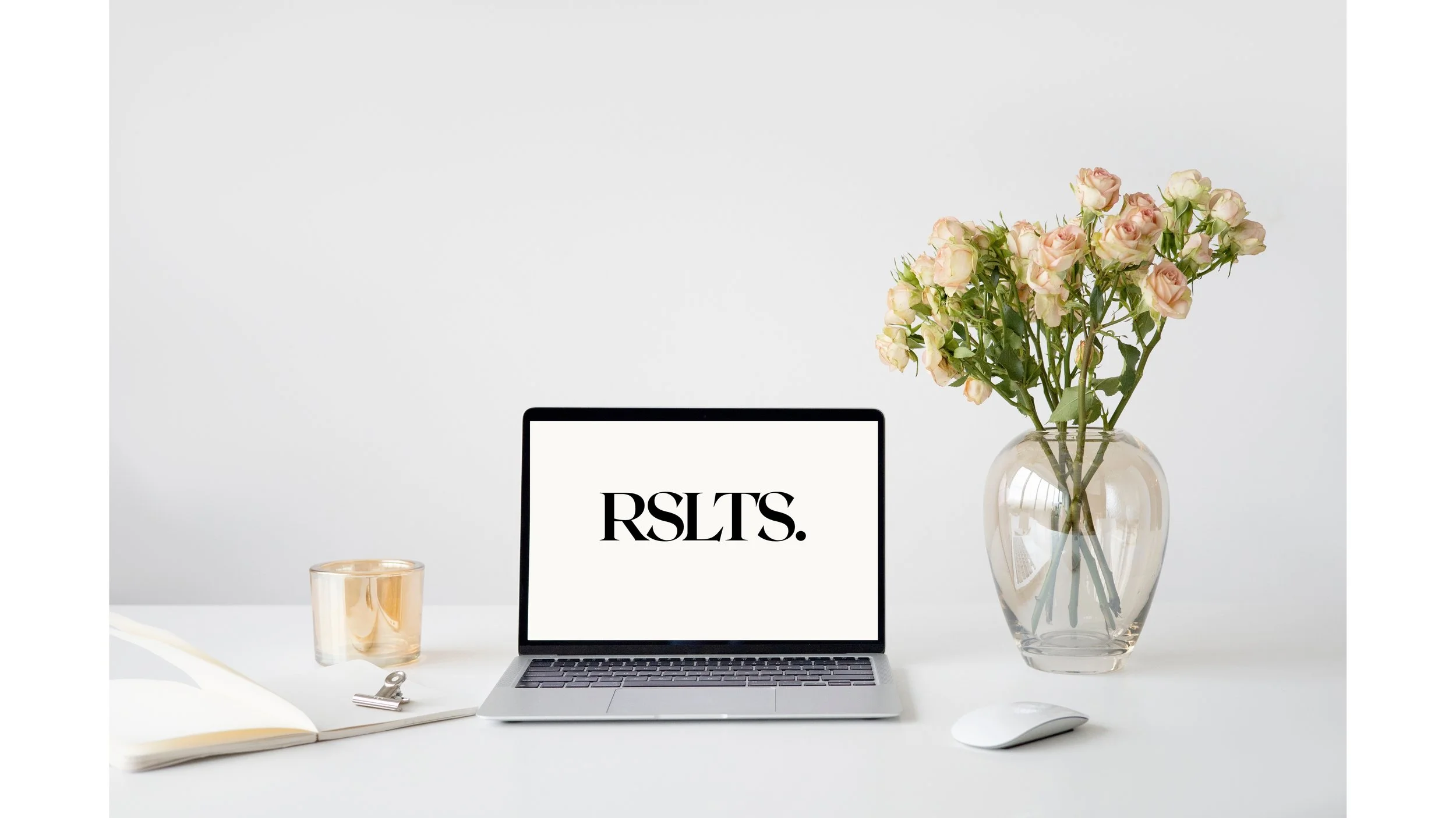 Minimalist workspace with a laptop displaying 'RSLTS', a mouse, a notebook with a clip, a candle, and a vase of pink roses on a white desk.