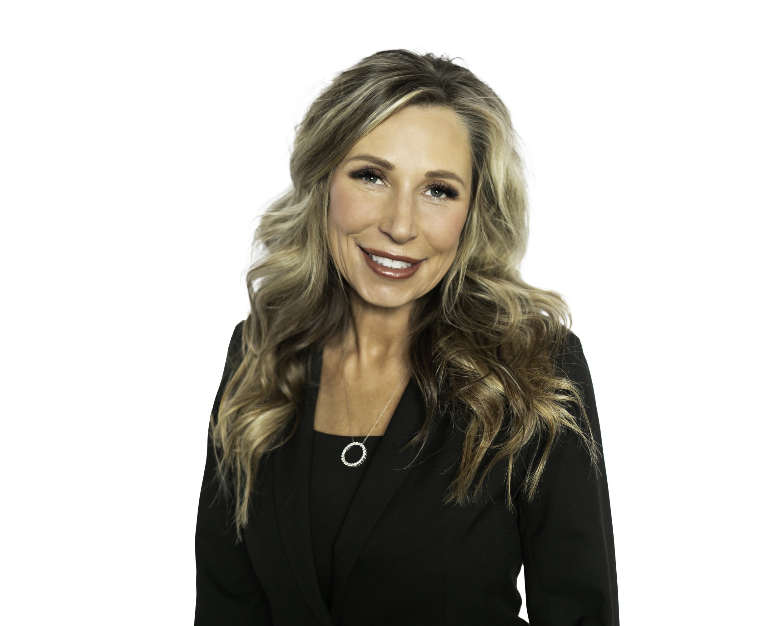 Portrait of a woman with long blonde hair wearing a black blazer and a circular pendant necklace, smiling against a white background.