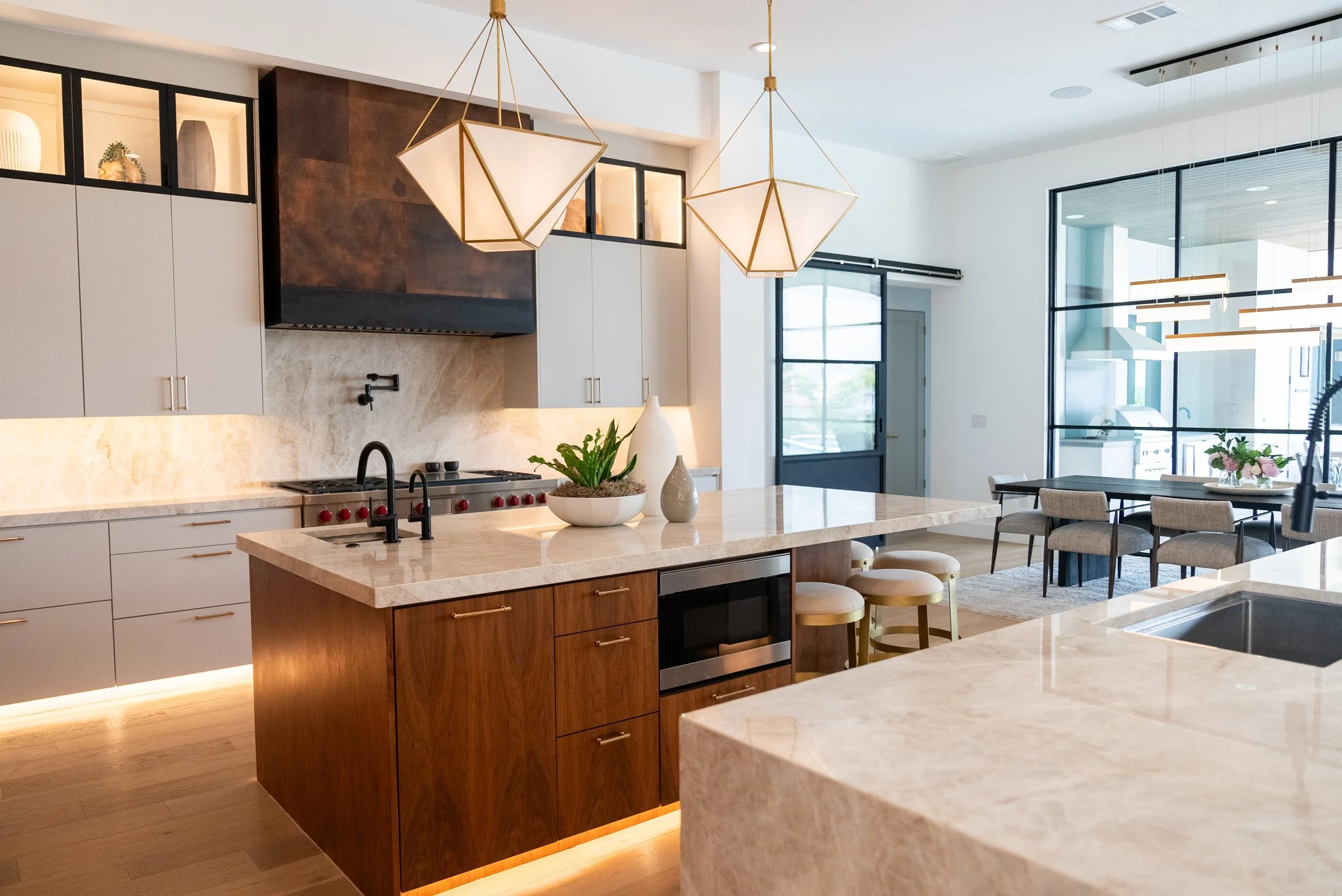 Modern kitchen with marble countertops, geometric pendant lights, white cabinets, dark stove hood, and dining area with a large window.