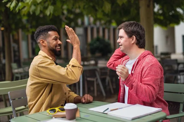 Two men sitting at a table outdoors, smiling and high-fiving each other, with notebooks and a coffee cup on the table.