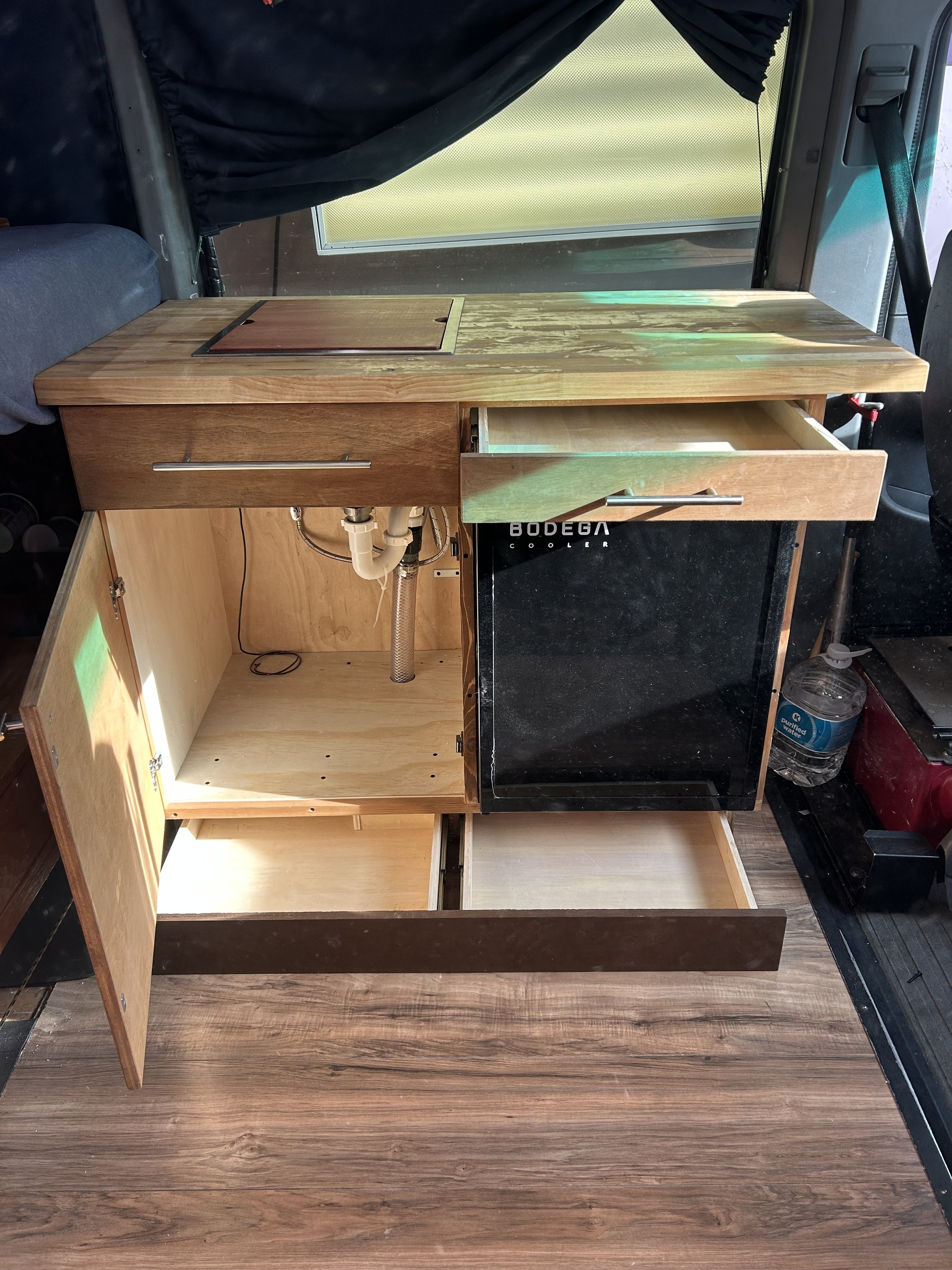 A kitchen galley added to a Sprinter T1N. Includes drop in sink and faucet with a cutting board to create maximum counter space, small drawer above the fridge, plenty of space under the sink and 2 hidden toe kick drawers. The toe kick drawers are wir