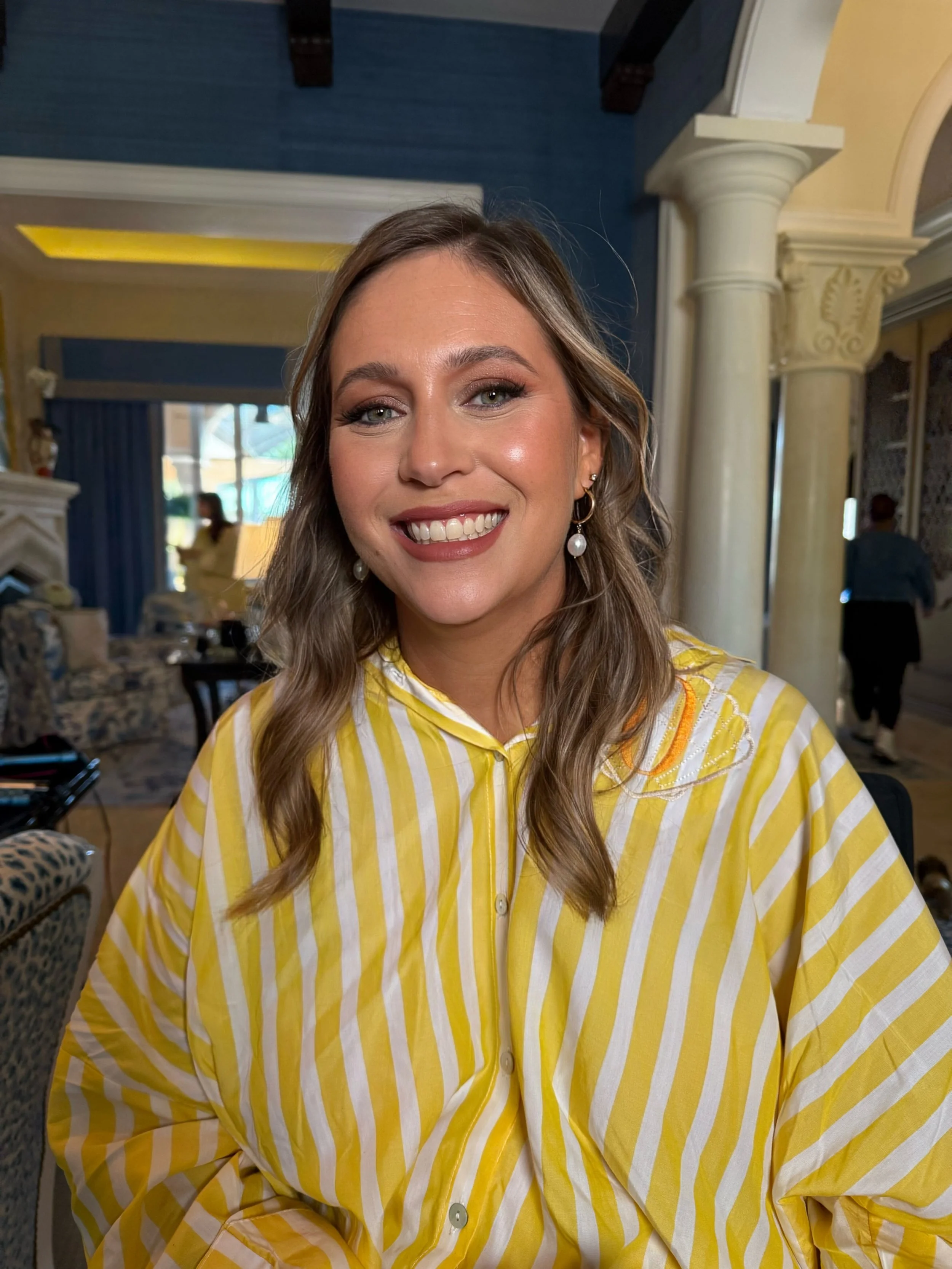 A woman with light brown hair, blue eyes, and pearl earrings, smiling, wearing a yellow and white striped shirt, in an indoor setting with classical decorative columns and a blue ceiling.