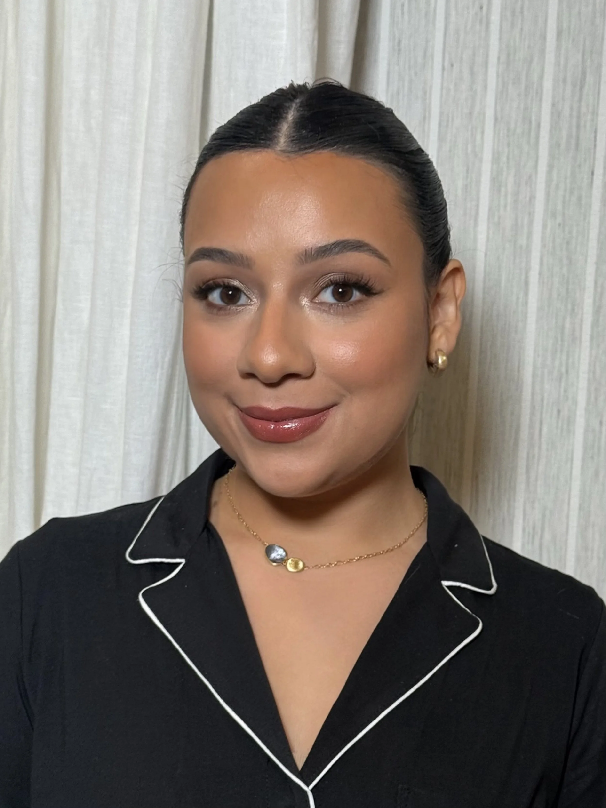 A woman with dark hair in a bun, wearing gold earrings, a layered gold necklace, and a black shirt with white piping. Behind her is a light-colored curtain.