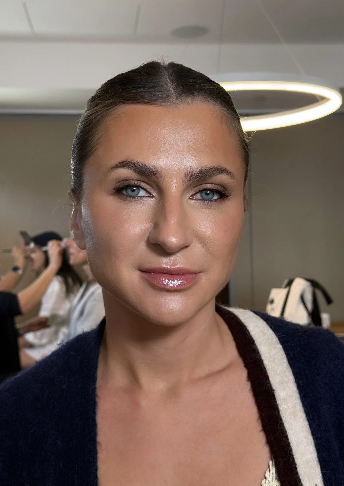 Close-up of a woman with pulled-back hair, wearing makeup, and a dark sweater with beige and dark trim. In the background, a person is applying makeup while another is taking a photo.