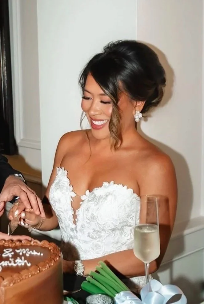 Woman in white strapless wedding dress smiling while cutting a cake, with champagne glass nearby.