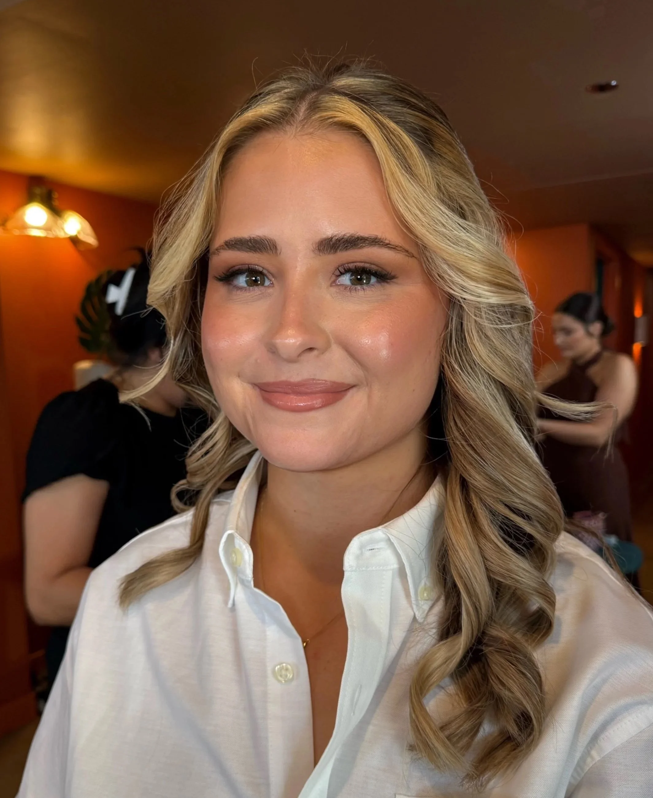 Close-up of a smiling woman with blonde, wavy hair, wearing a white shirt, in an indoor setting with warm lighting and a blurred background.